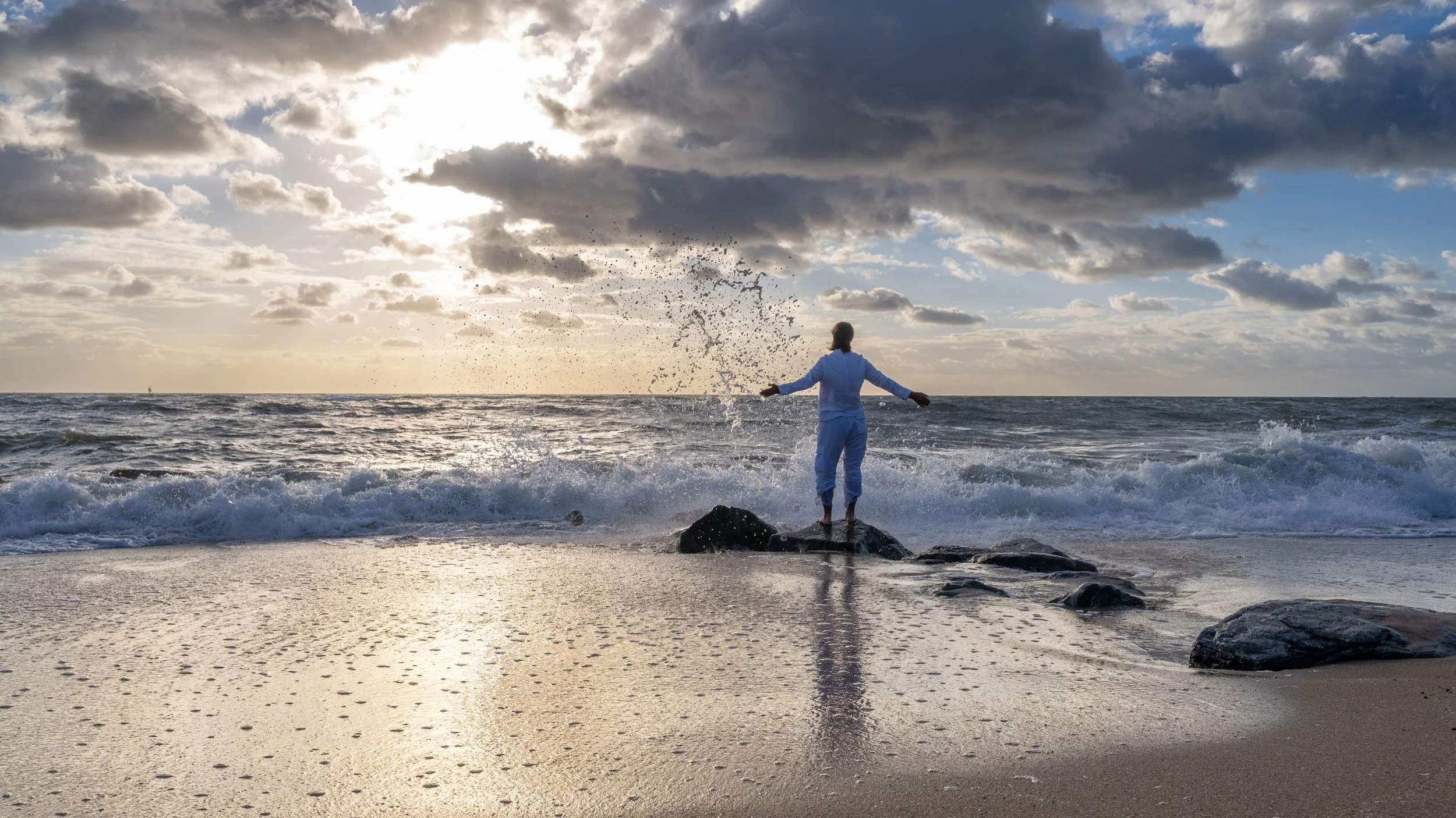 Person standing on rocks at the beach during sunset, with waves crashing and cloudy sky in the background.