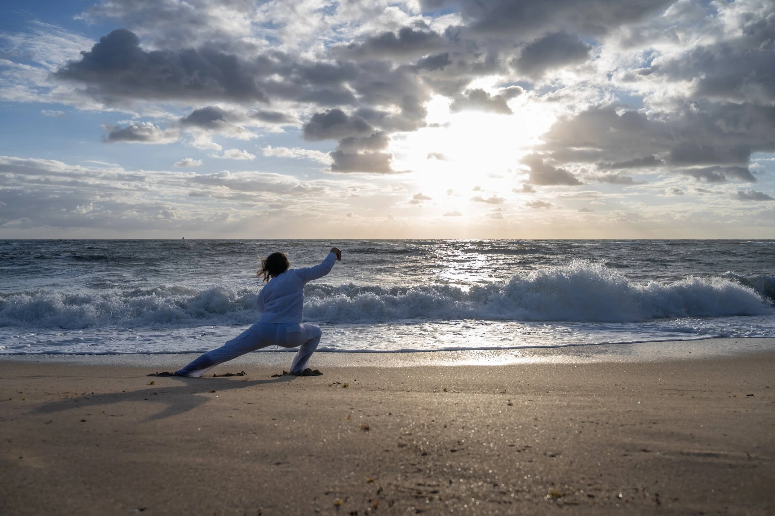 Person in white clothing practicing yoga on a sandy beach during sunset, with ocean waves and a cloudy sky in the background.