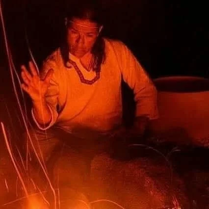 A woman dressed in a light-colored top, sitting near a fire with sparks flying, in a dark setting.