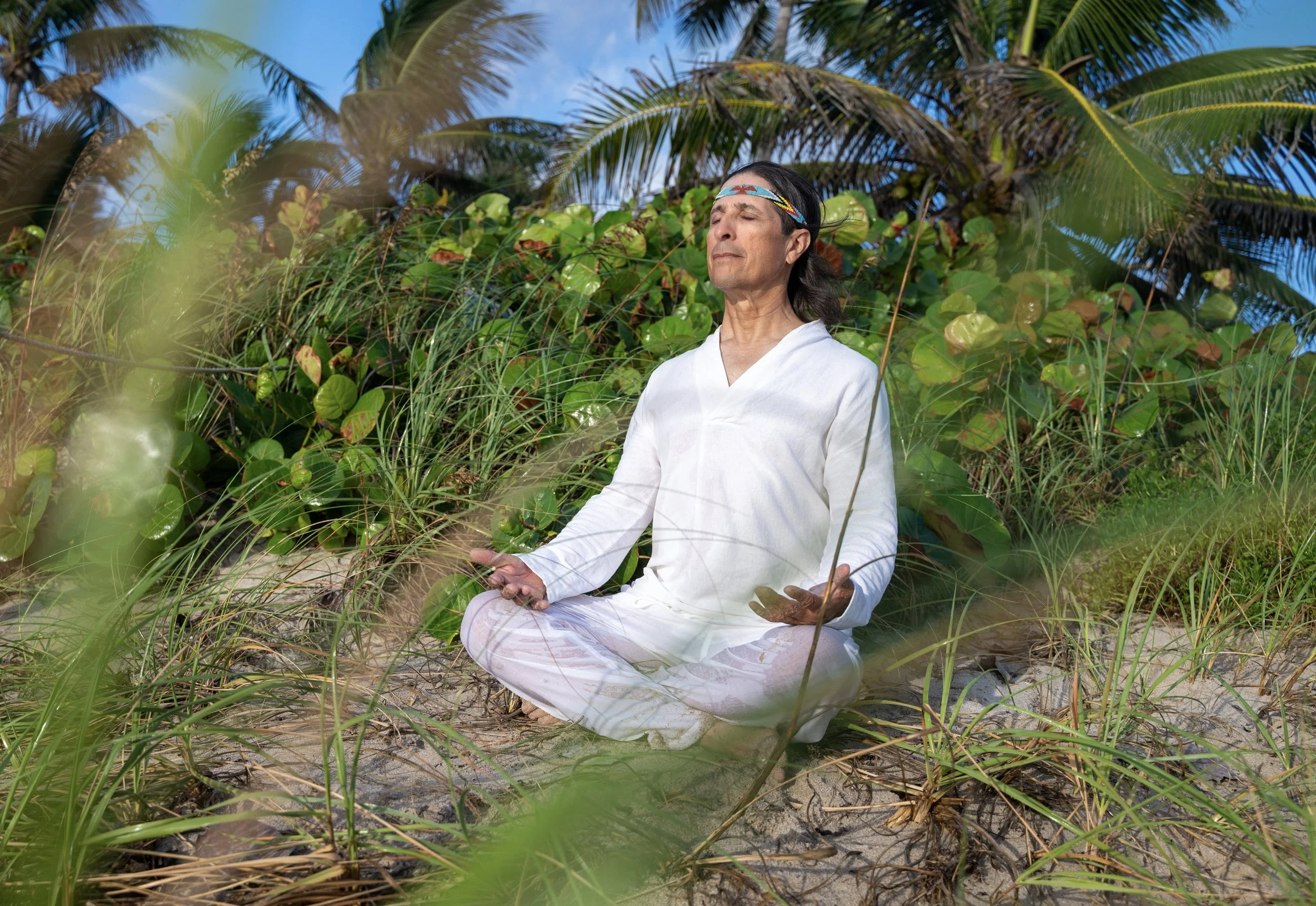 A person practicing yoga or meditation outdoors on a sandy surface surrounded by green foliage and palm trees, with eyes closed and a peaceful expression.