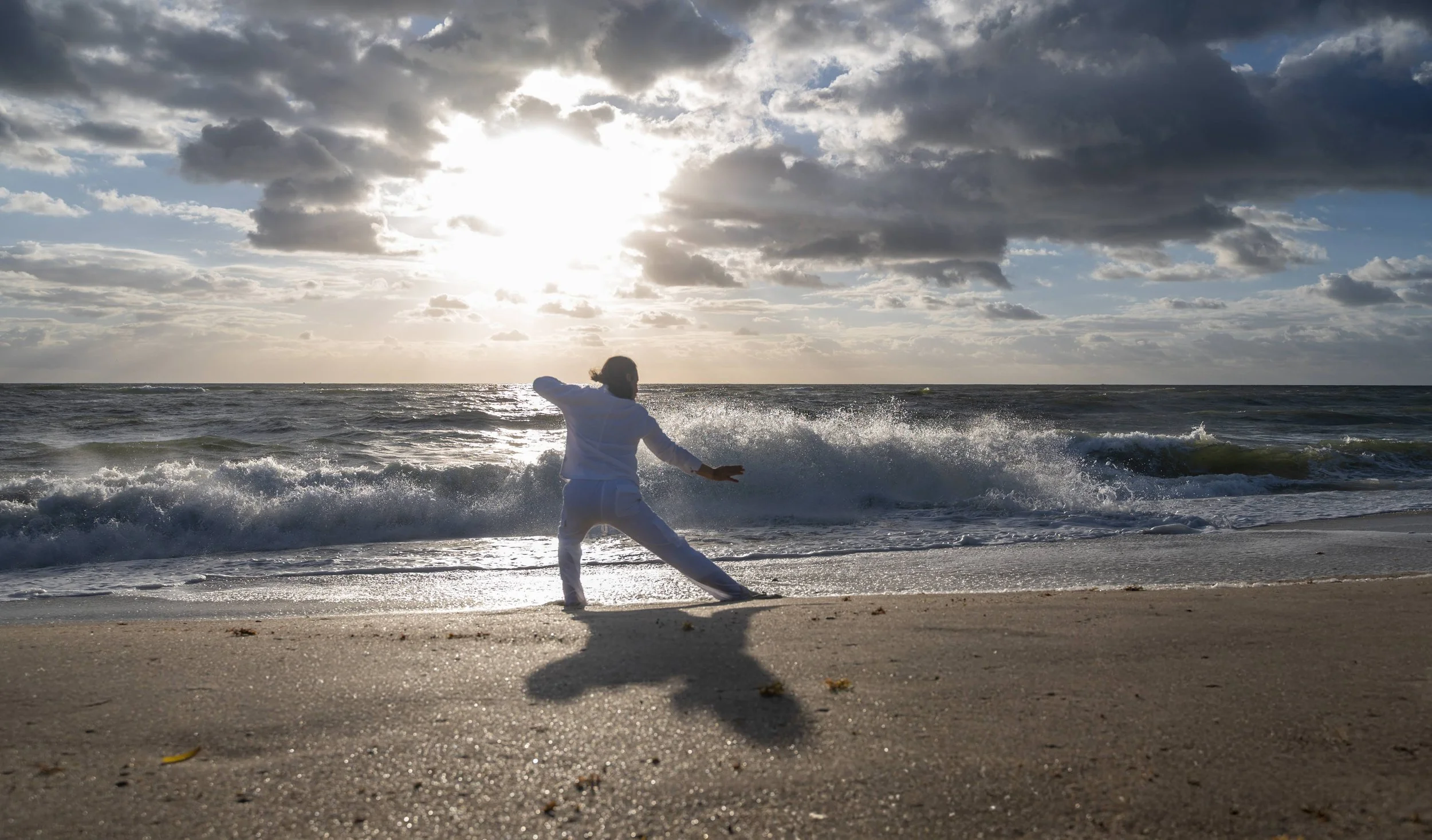 Person in white attire practicing a martial arts move on the beach at sunset, with waves crashing nearby and cloudy sky overhead.
