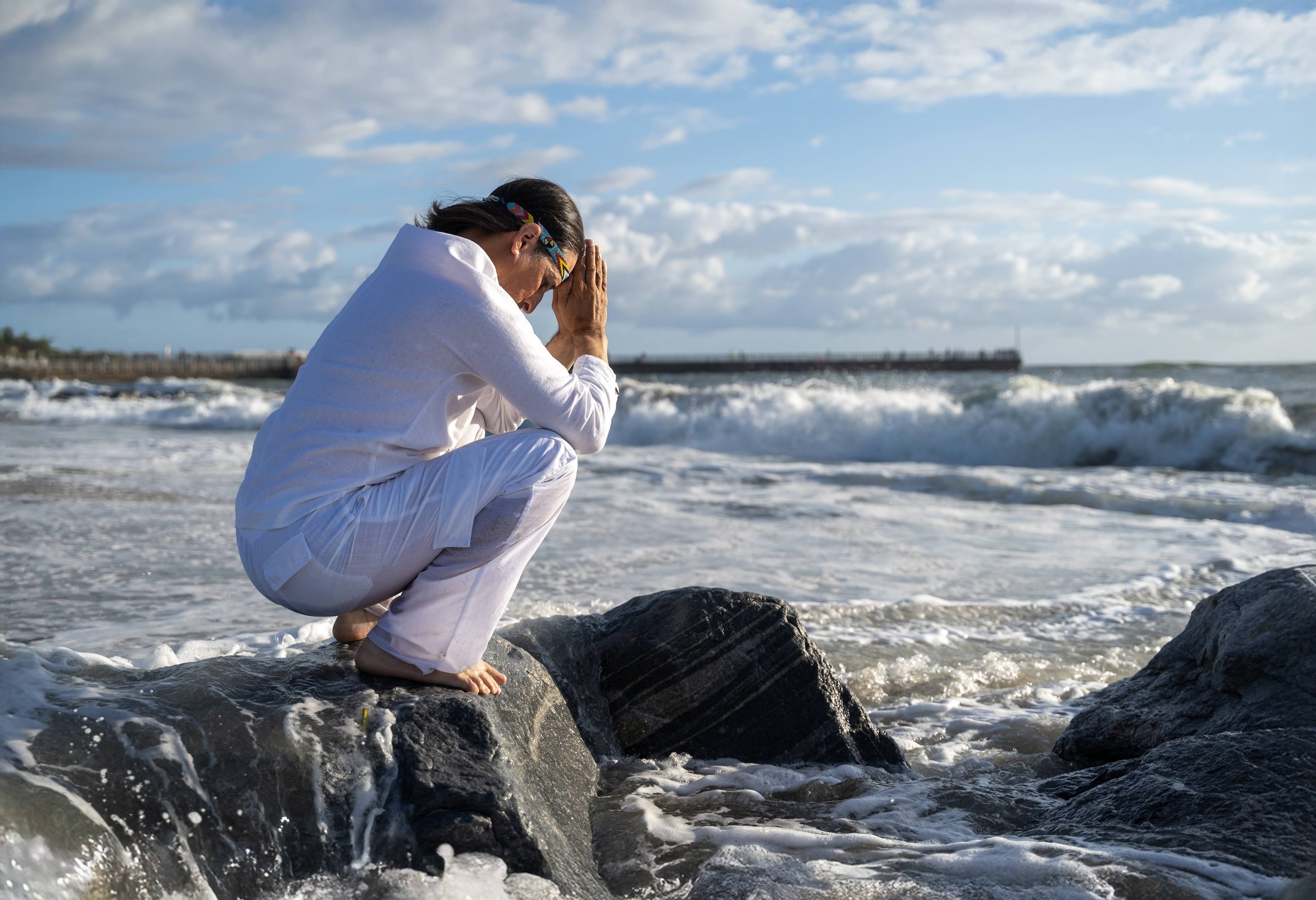 Person kneeling on rocks at the beach with hands in prayer position, wearing white clothing and a colorful headband, facing the ocean with waves and a cloudy sky in the background.