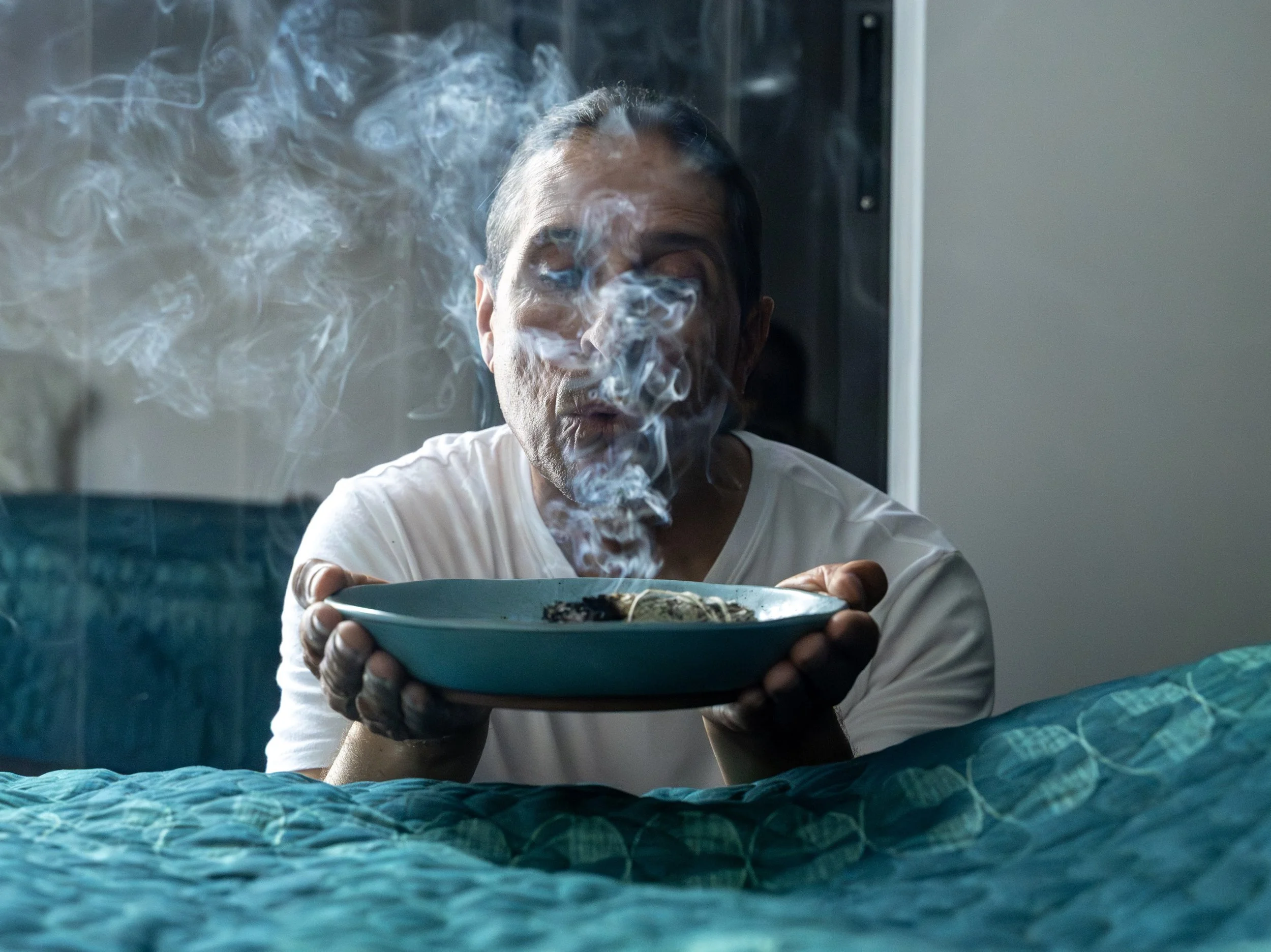 A man with salt-and-pepper hair and light skin, wearing a white shirt, is sitting on a bed and holding a bowl of steaming hot food. Smoke or steam is rising from the bowl, obscuring part of his face as he leans forward to blow on the food.