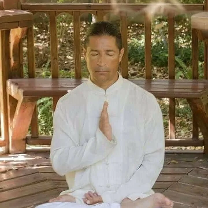 A man in white clothing practicing meditation or yoga outdoors on a wooden deck, with his eyes closed and hands in a prayer position.