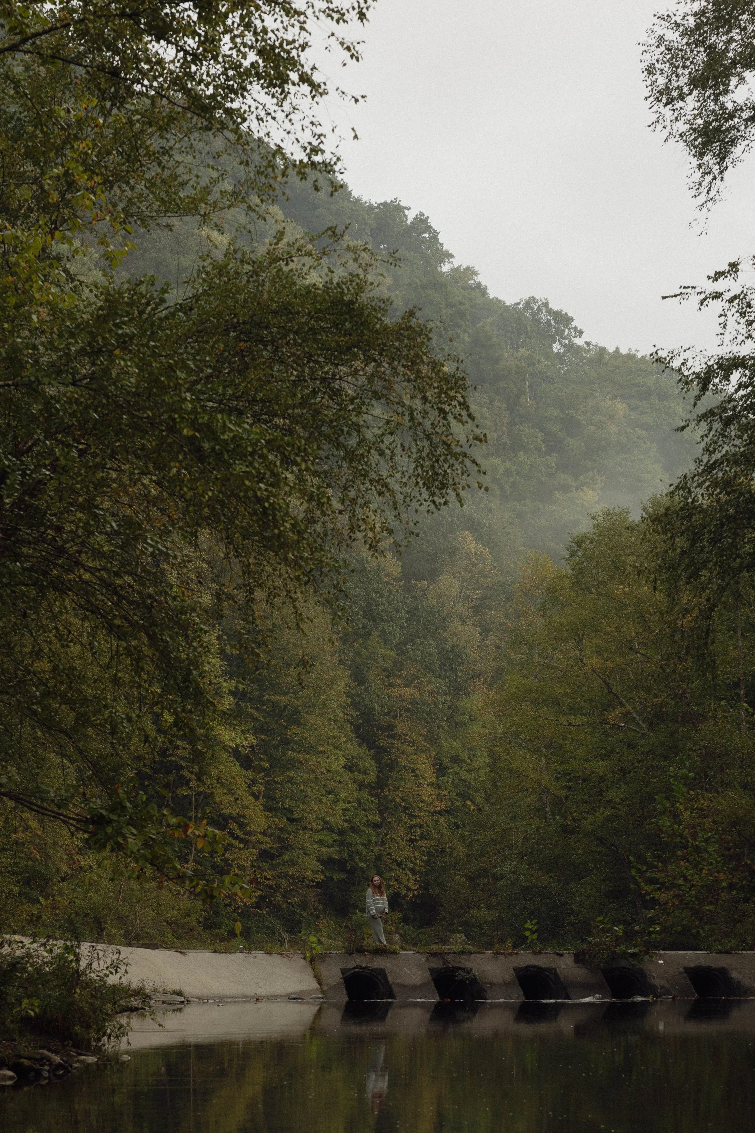 A woman standing on a concrete structure by a river in a lush green forest with overcast sky