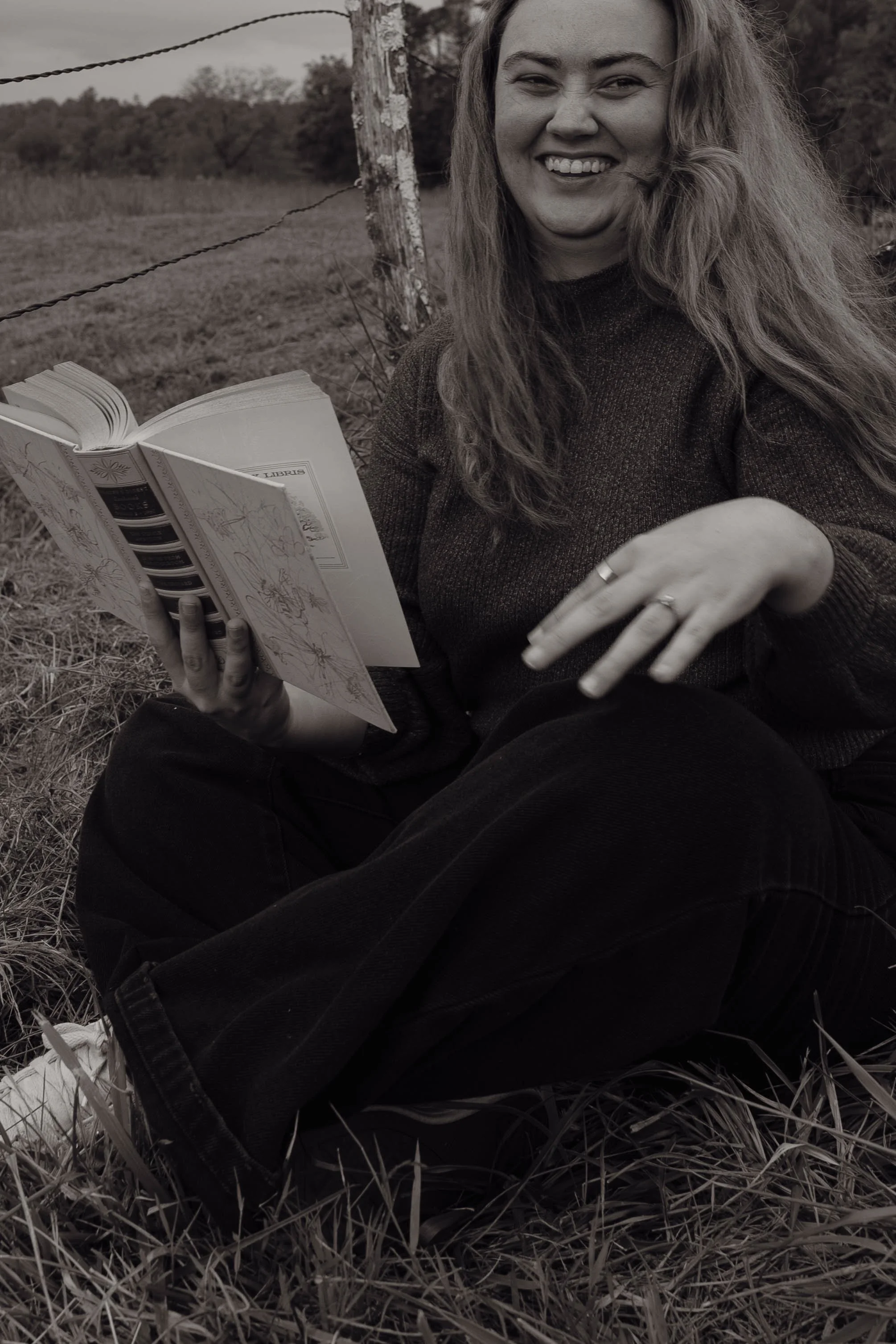 A woman sitting on the grass outdoors, smiling, holding an open book, with a fence and trees in the background.