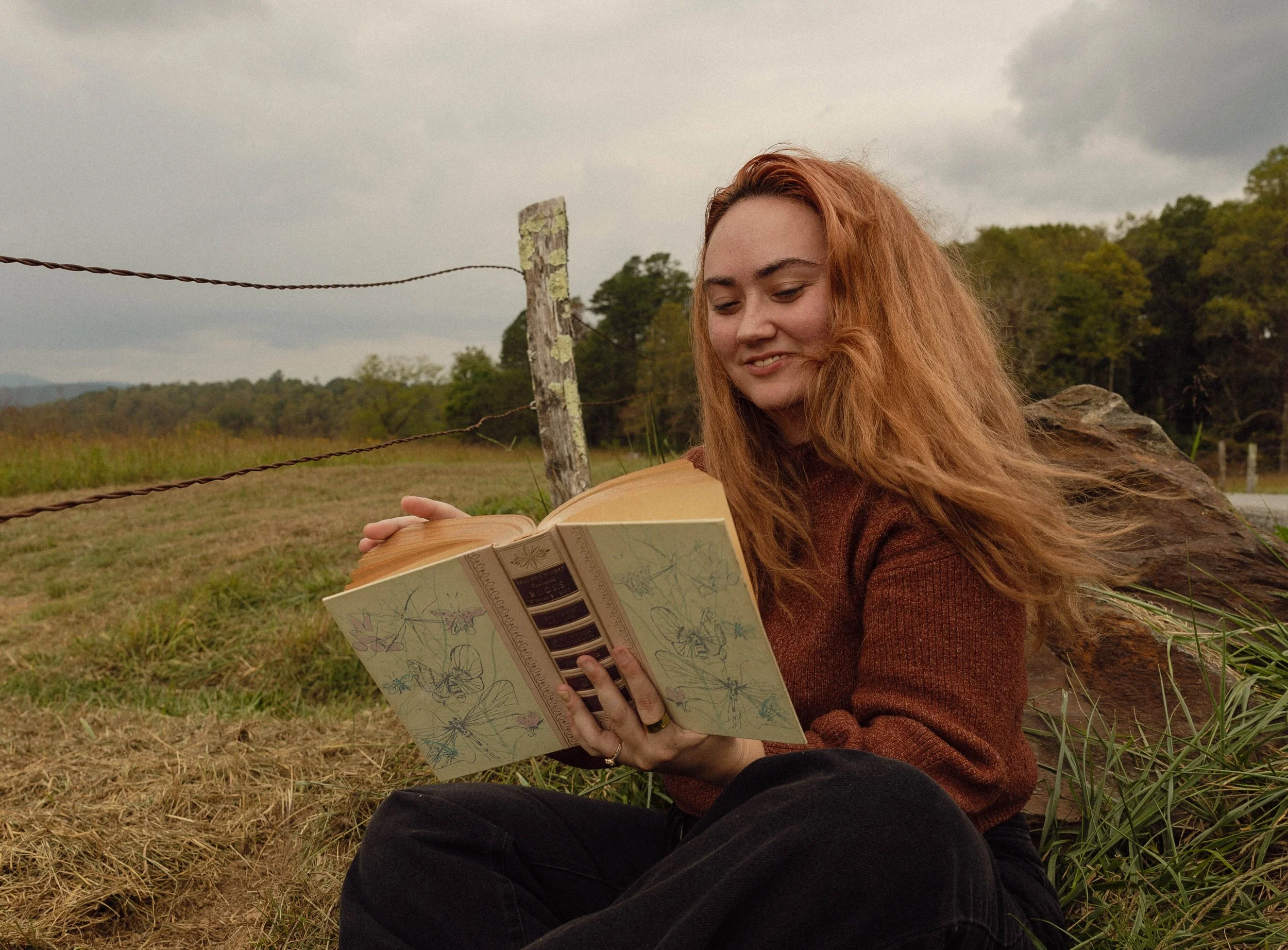 A young woman with red hair reading a book outdoors near a fence and trees.