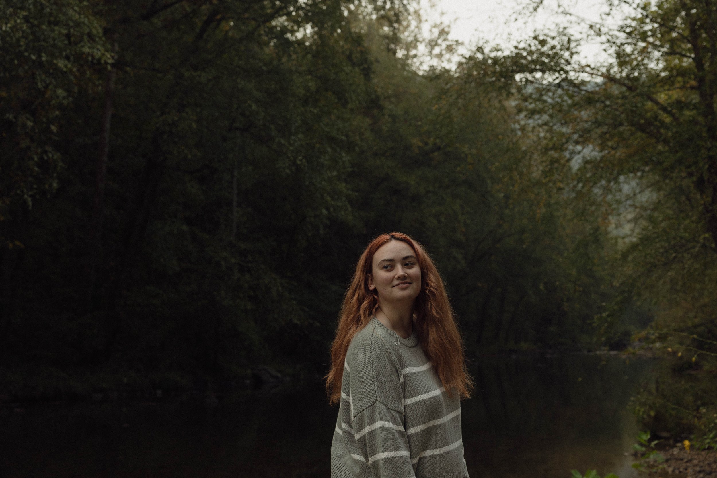 A young woman with long red hair standing outdoors near a calm river surrounded by trees in a forest.