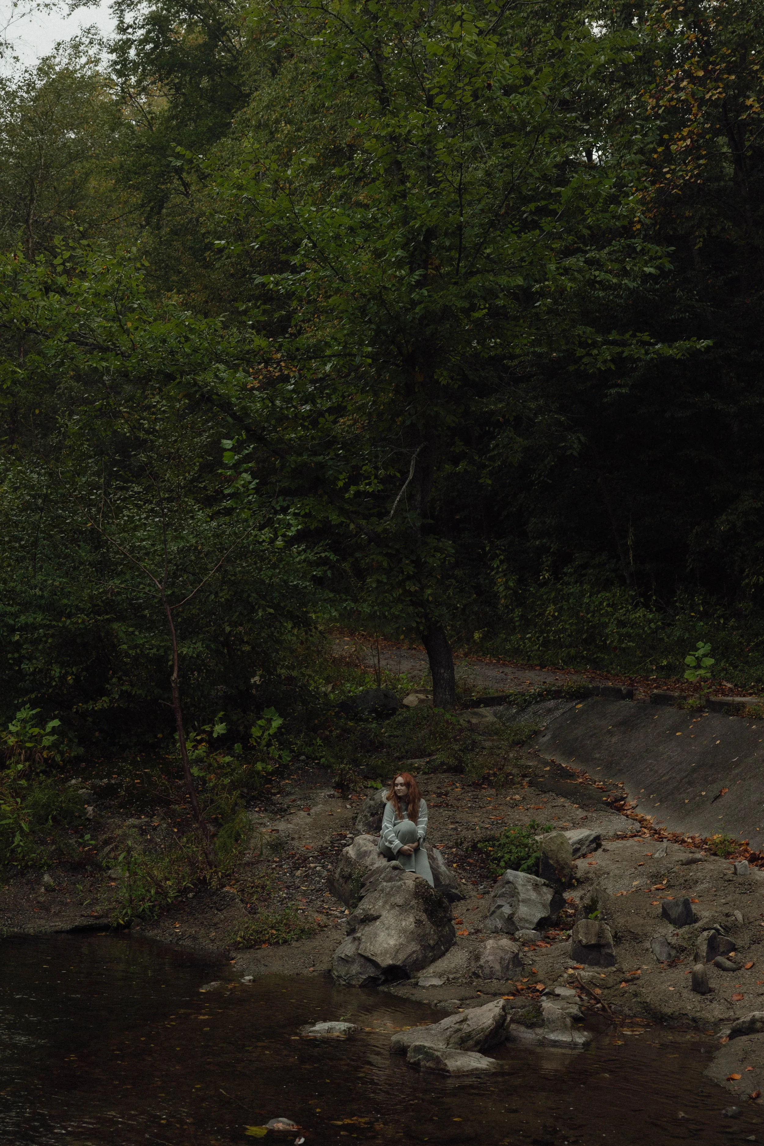 A woman with long red hair sits on a large rock near a small body of water, surrounded by green trees in a forested area during autumn.