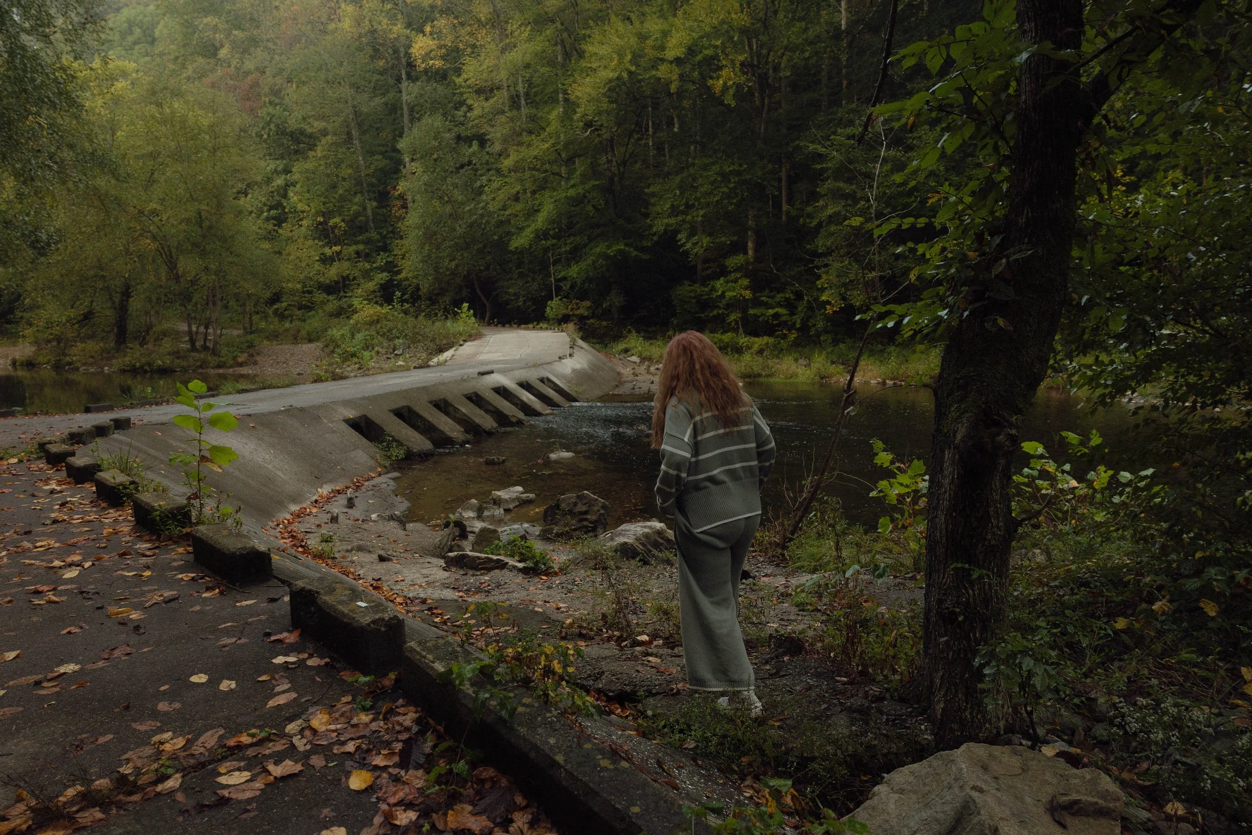 A person with long, curly hair walks along a path next to a small river in a wooded area during autumn. The path is covered with fallen leaves, and the water level appears high, partially flooding the path.