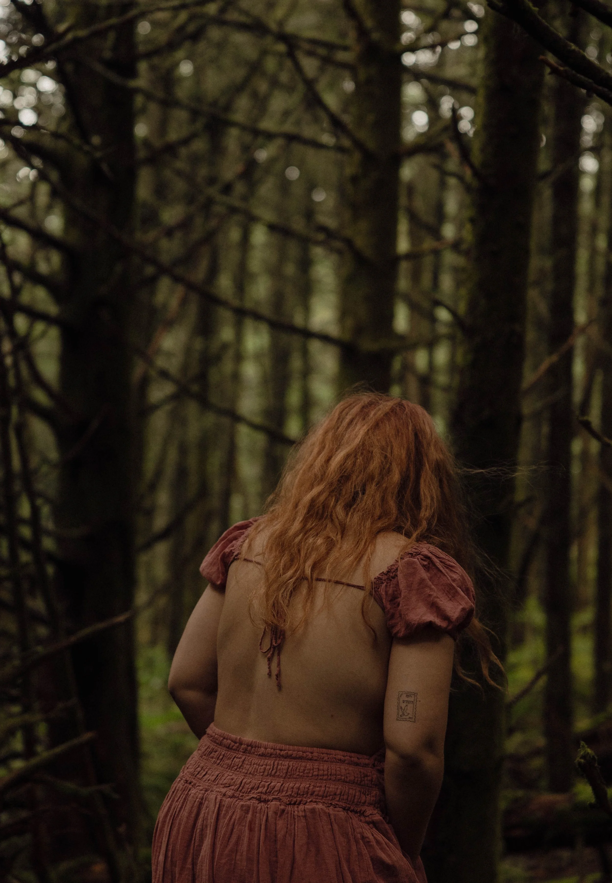 A woman with long red hair wearing a beige top with puffed sleeves and a long, high-waisted rust-colored skirt, standing in a dense forest with tall trees and green foliage.