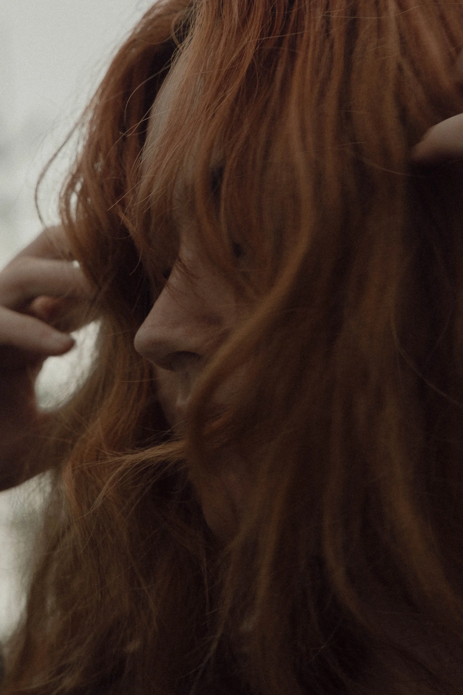Close-up of a woman with long, wavy red hair, holding a section of her hair with one hand and looking downward.