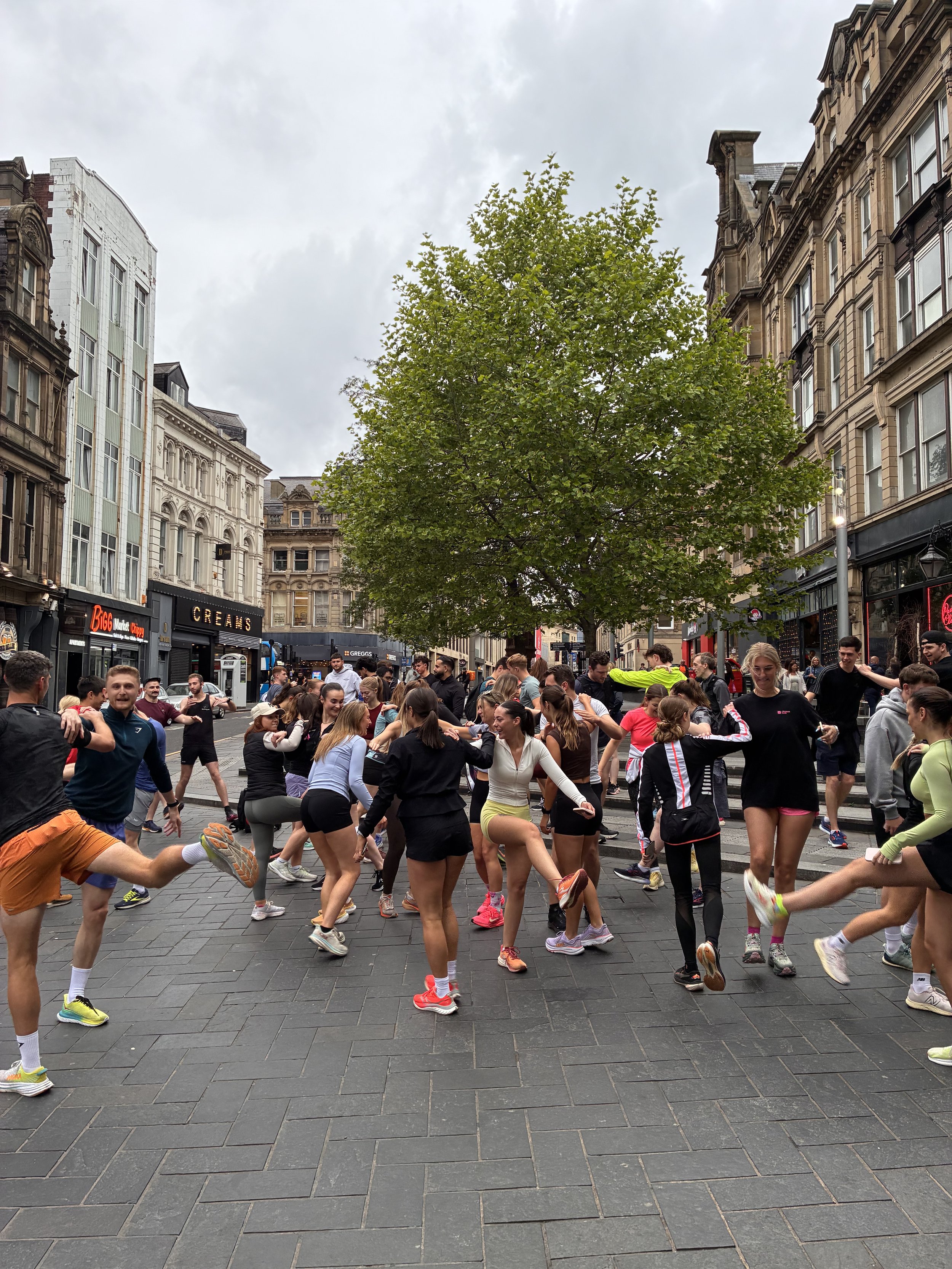 Group of people doing an outdoor workout or dance routine on a city street with tall buildings and a green tree in the background.