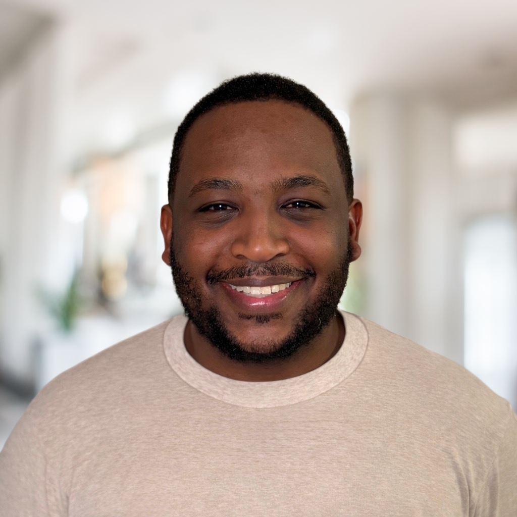 A smiling Black man with short hair and a beard, wearing a beige t-shirt, standing in a bright room with blurred background.