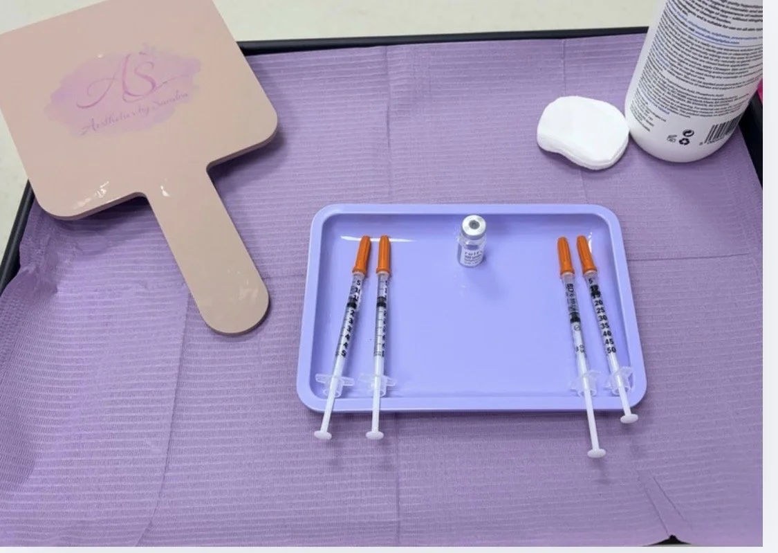 Syringes, alcohol wipe, and a small bottle on a purple tray, with a cotton pad, a bottle, and a mirror with a pink handle on a purple table.