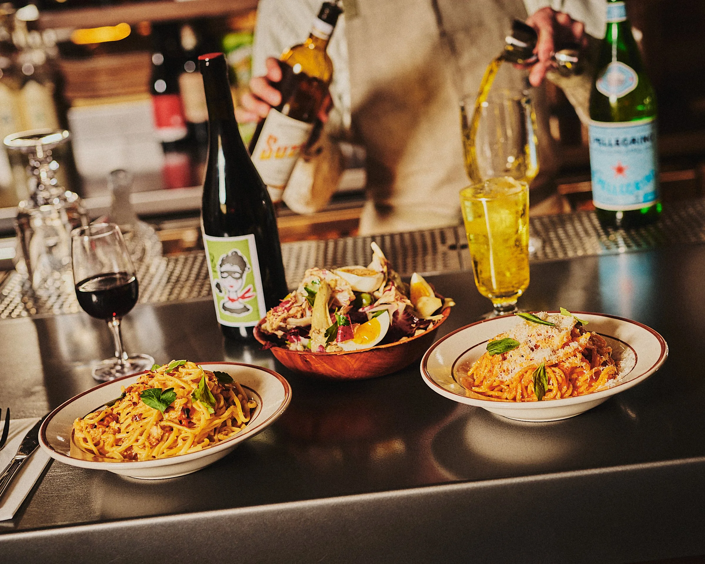 A bar counter with plates of pasta, a salad, a glass of red wine, a beer bottle, and two glasses of beer, with a bartender pouring a drink in the background.