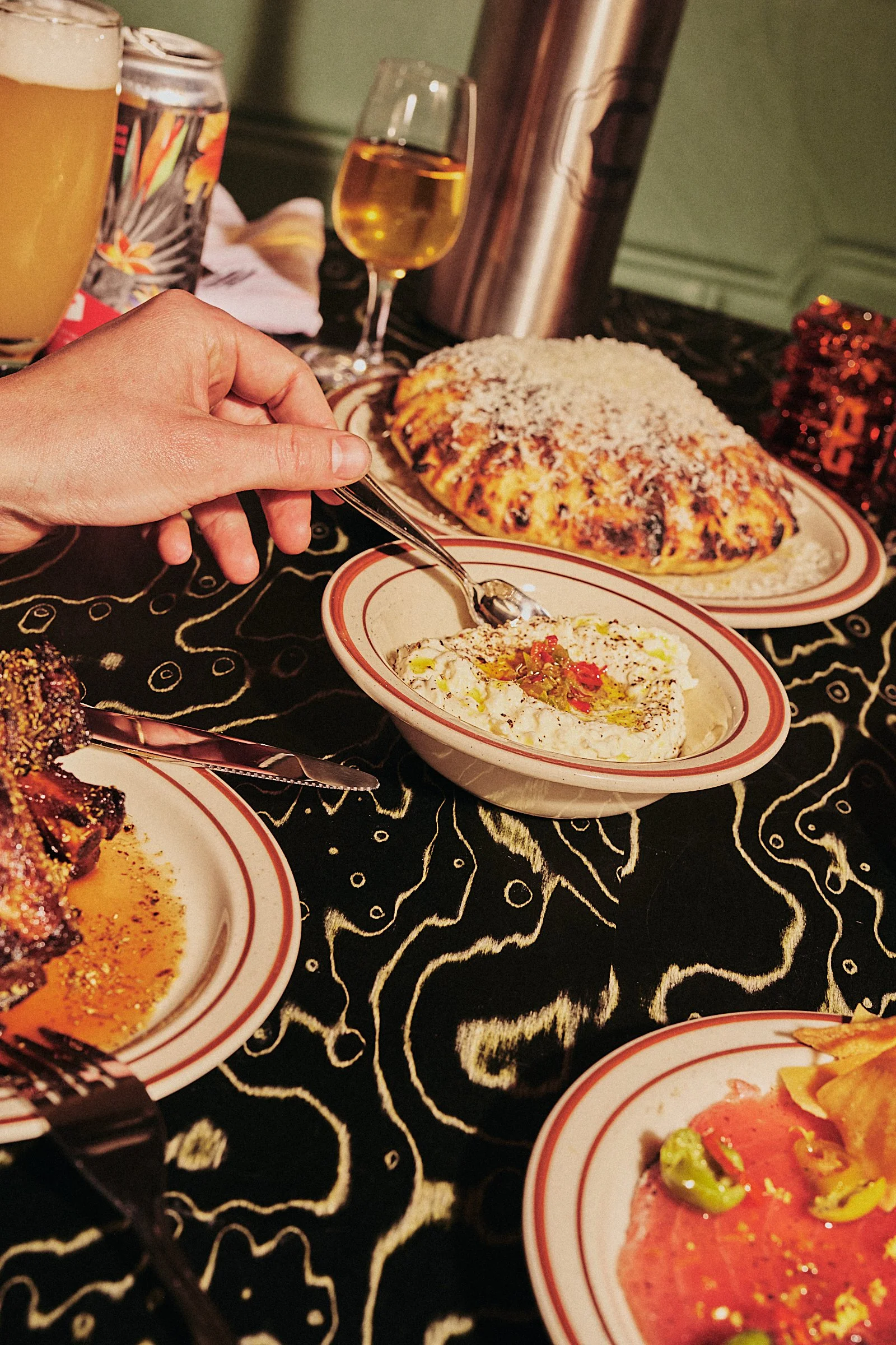 A table with various dishes including a large rolled casserole or stuffed bread topped with cheese, a bowl of creamy dip garnished with red and green peppers, a plate of sliced cured meat with peppers, and a partially visible dish with sauce and garn