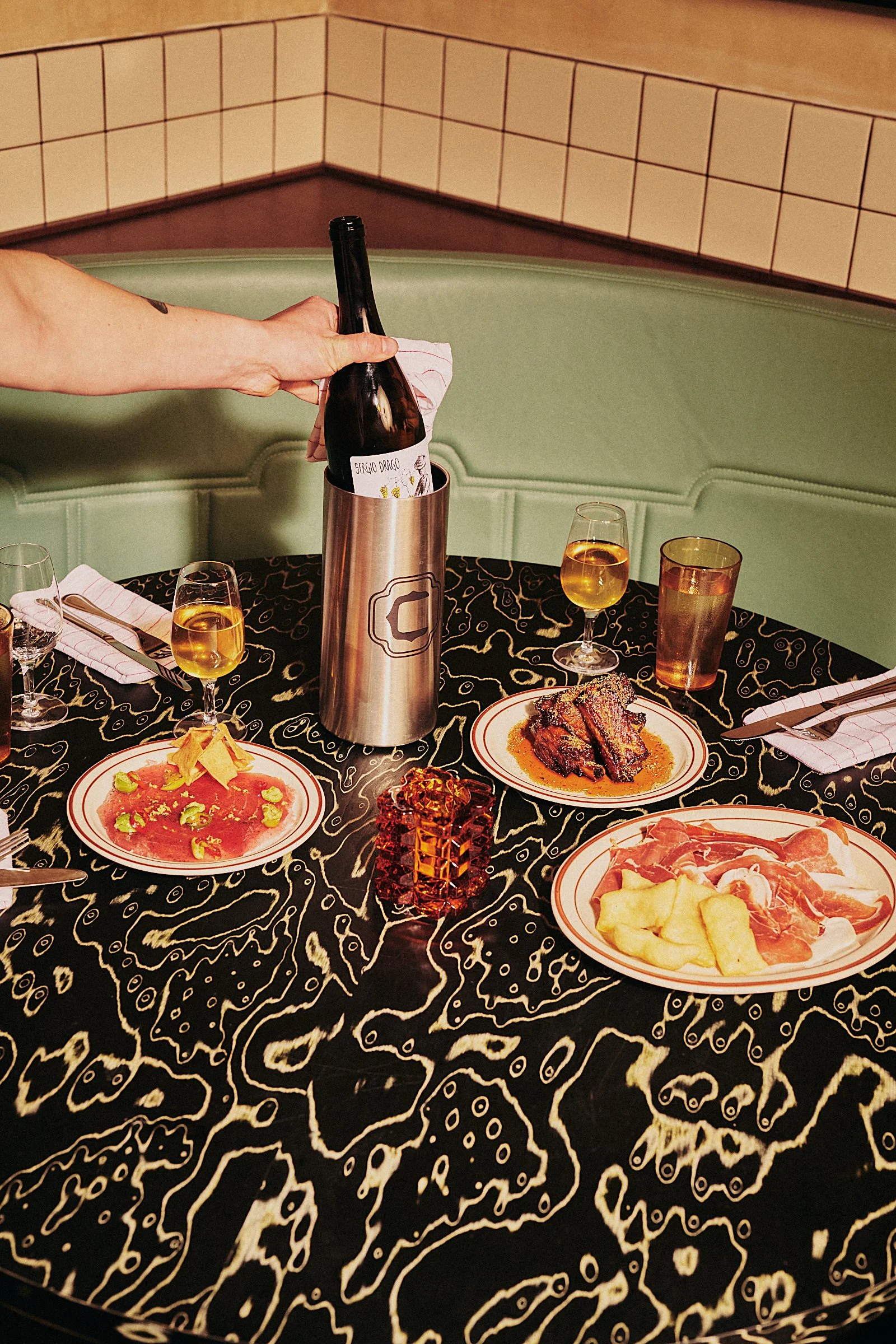 A table set for a meal with plates of raw meat, cooked meat, and chips, glasses of white wine, a large bottle of wine in a silver wine cooler, and a small orange decorative block, in a restaurant with tiled walls and green seating.