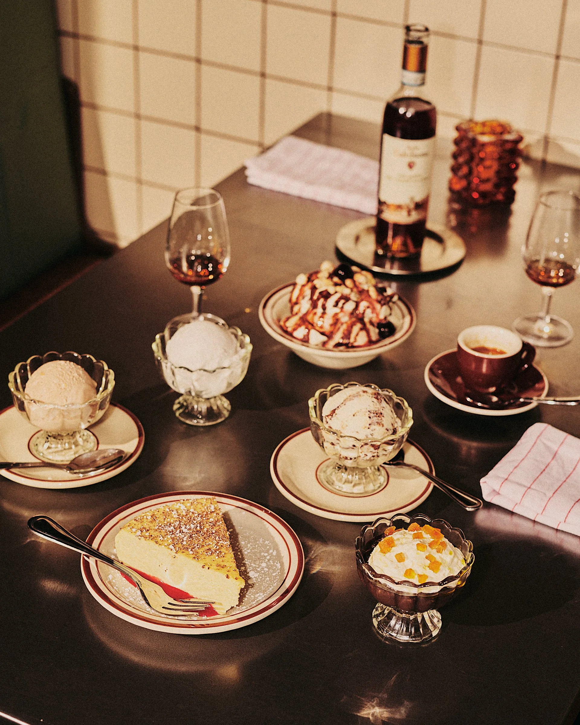 A table set with several cups of coffee, glasses of wine, bowls of ice cream, a slice of cheesecake, and a dessert topped with syrup and fruit, with a bottle of wine in the background.