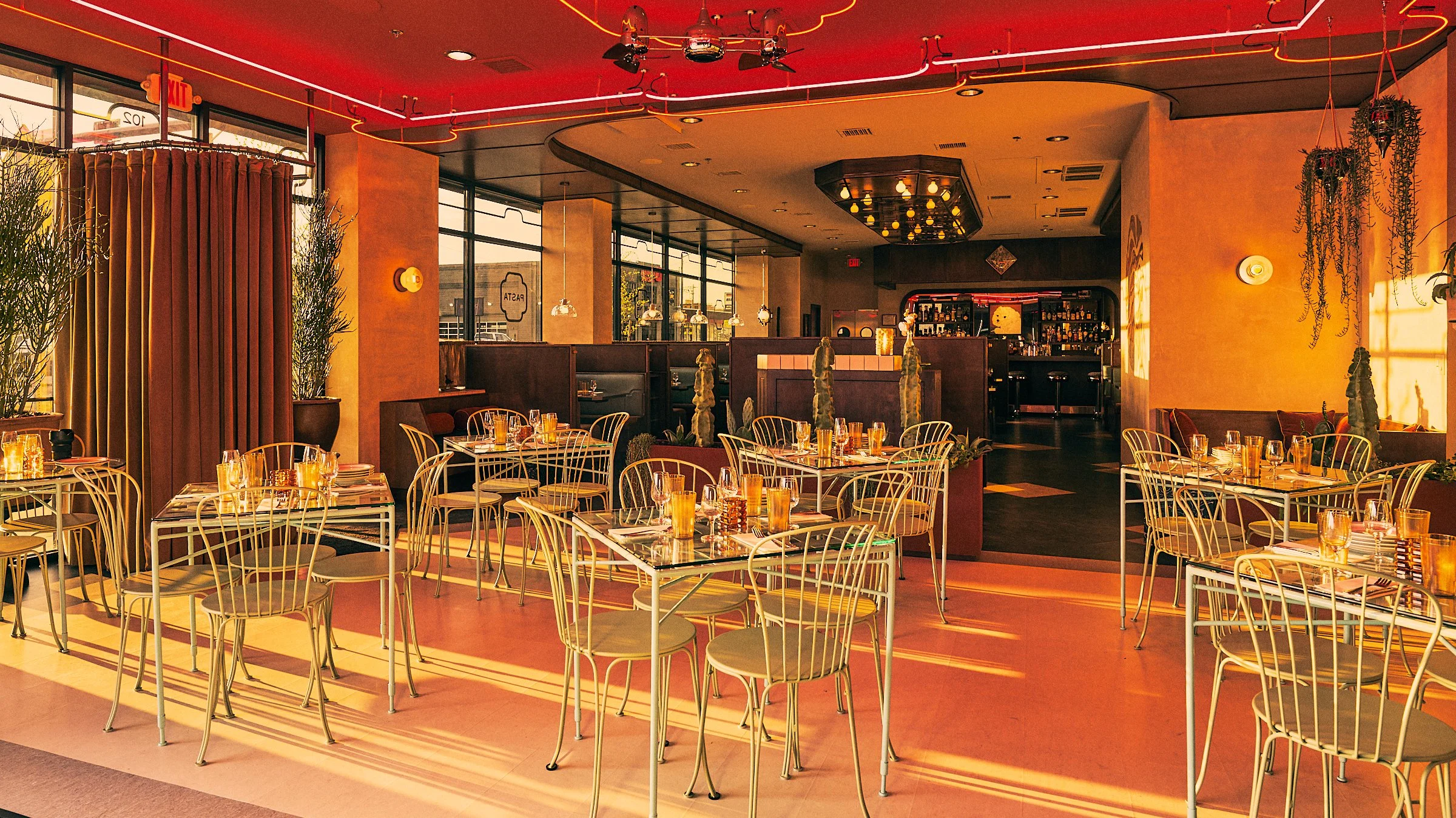 Empty restaurant with metal chairs and tables set with glasses and napkins, sunlight streaming through large windows, warm ambient lighting, decor includes plants and hanging lights, with a bar area in the background.