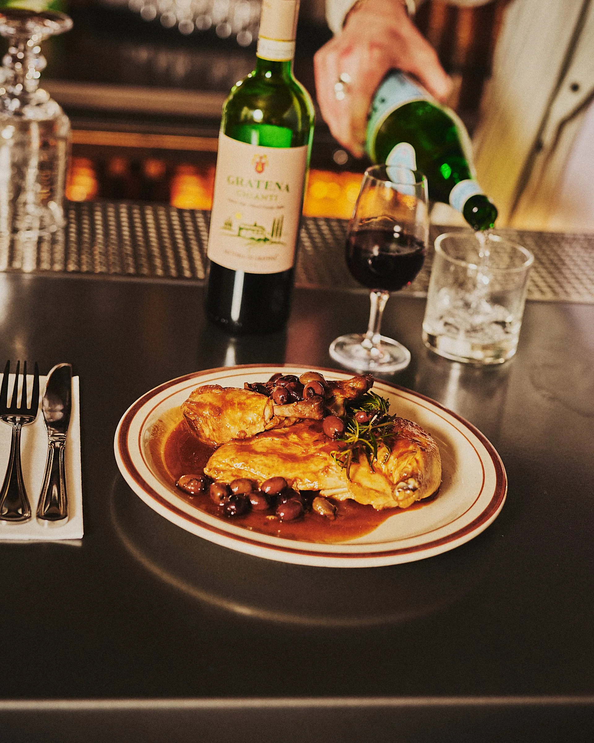A plated serving of chicken with sauce and olives, a glass of red wine, and a bottle of wine being poured at a bar.