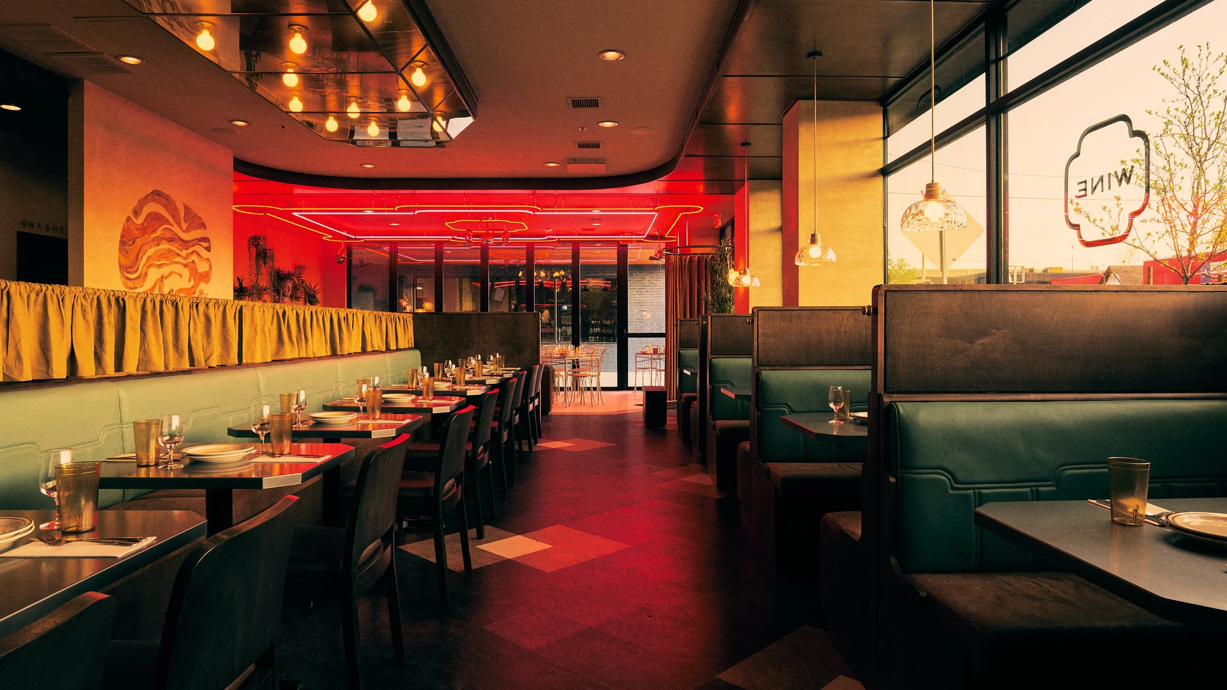 Empty restaurant with booths and tables, decorated with hanging pendant lights and neon ceiling lights, large windows with sunlight, and wall art inside.
