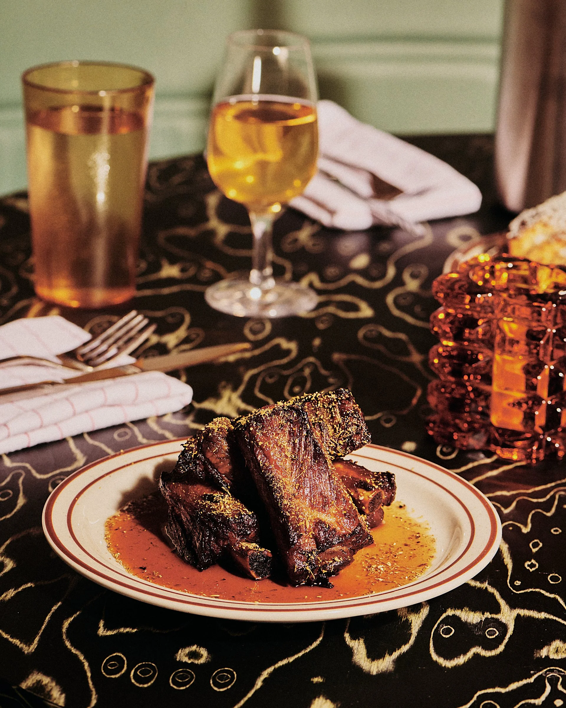 A plate of cooked beef short ribs on a table with a glass of white wine, a tall glass of a beverage, a fork and napkin, and a decorative glass sculpture.