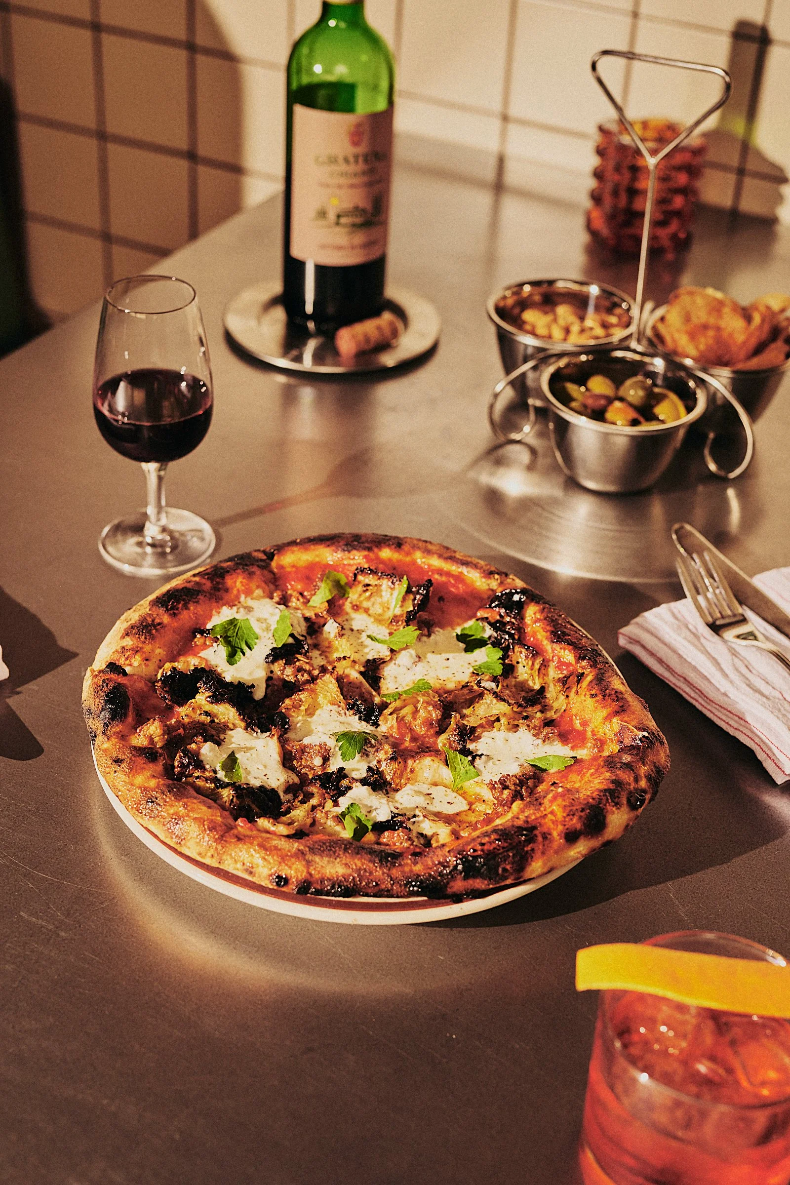 A pizza with toppings like cheese, black olives, and herbs on a dark table with a glass of red wine, a bottle of wine, a bowl of mixed olives, and bowls of breaded snacks.