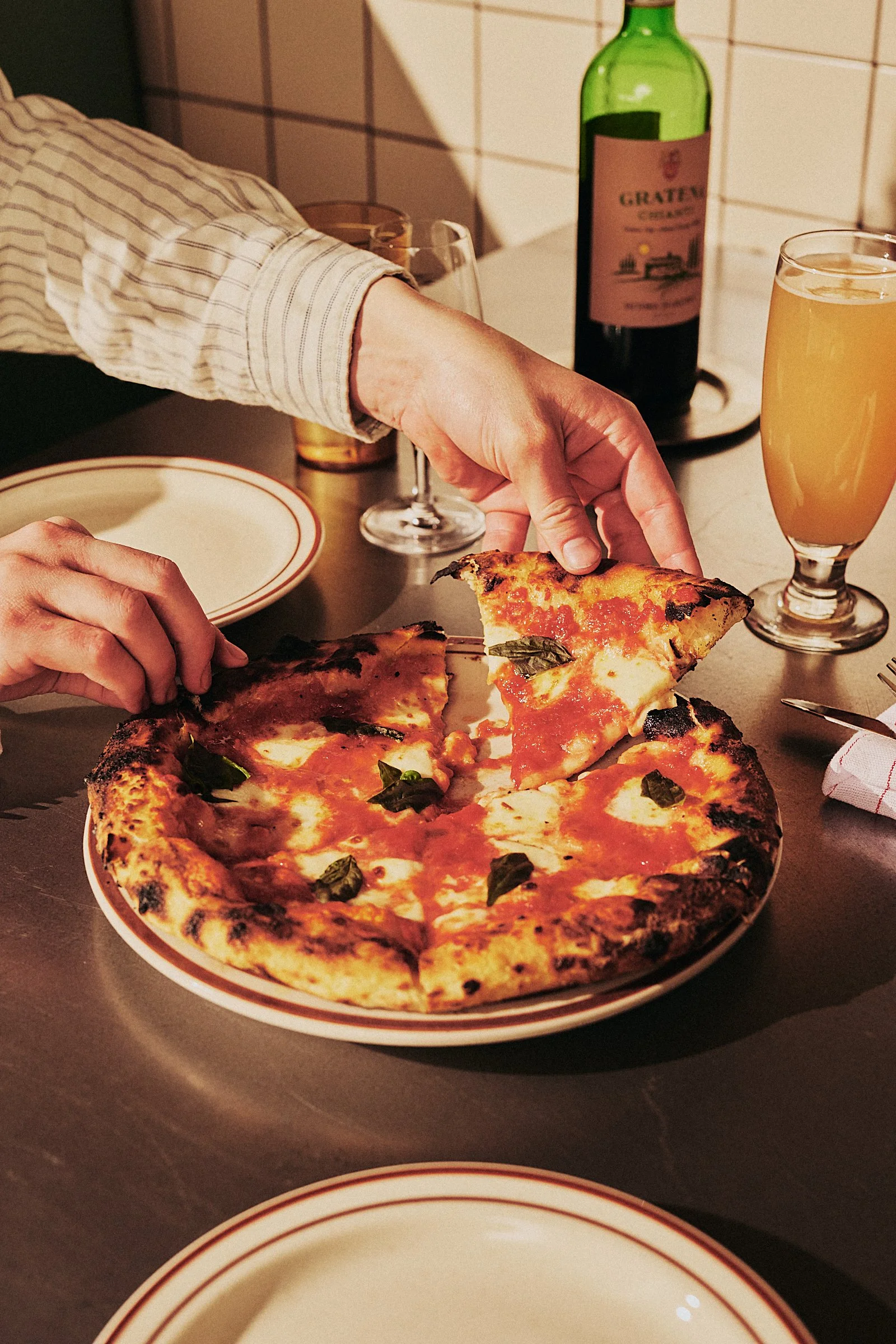 Person scooping up a slice of pizza from a plate that has several slices remaining, with a bottle of wine and two glasses on the table.