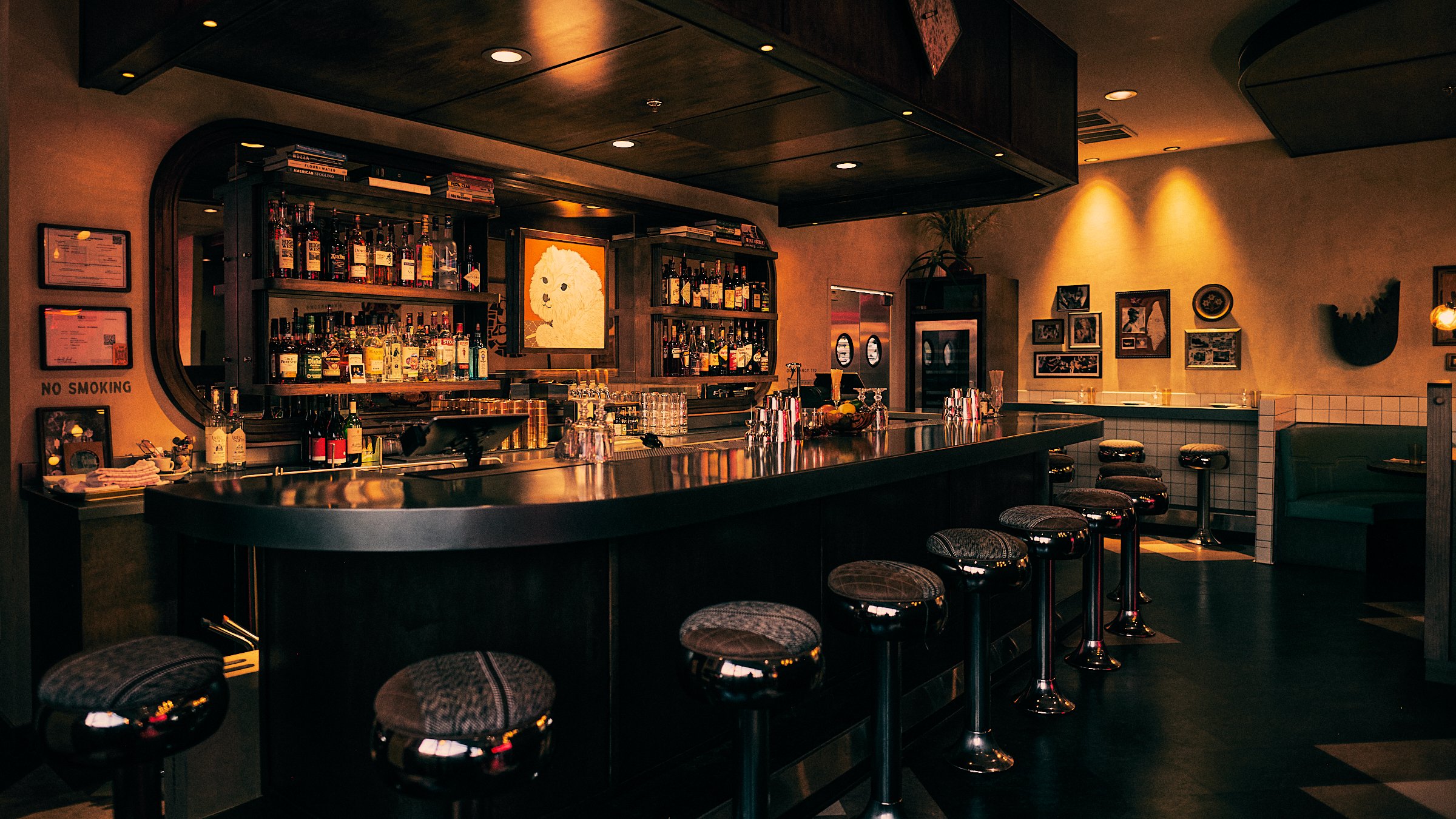 Interior of a dimly lit cozy bar with a curved counter, bar stools, shelves stocked with liquor bottles, framed pictures on the wall, and warm lighting.