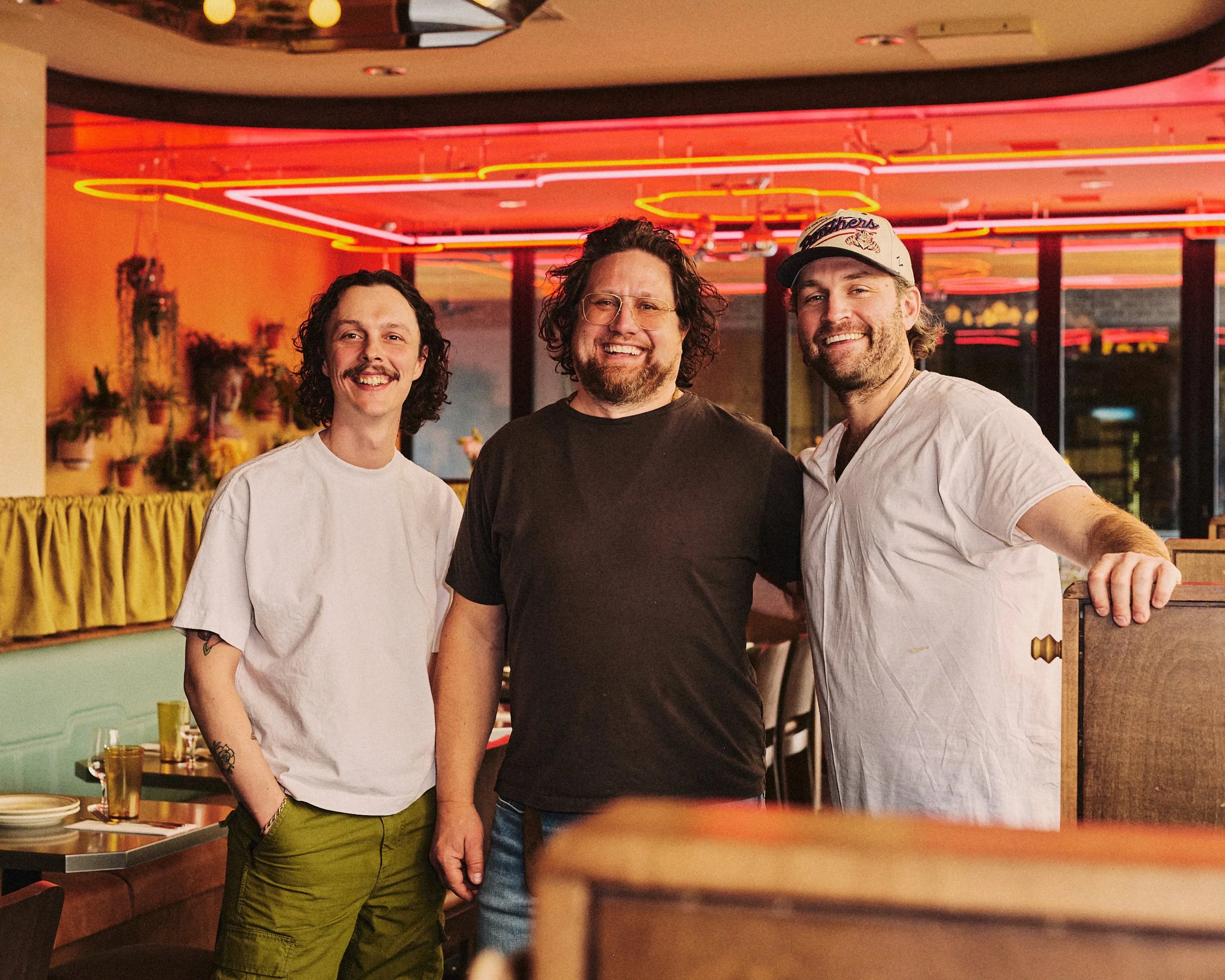Three men smiling in a restaurant with colorful neon lighting and plants in the background.