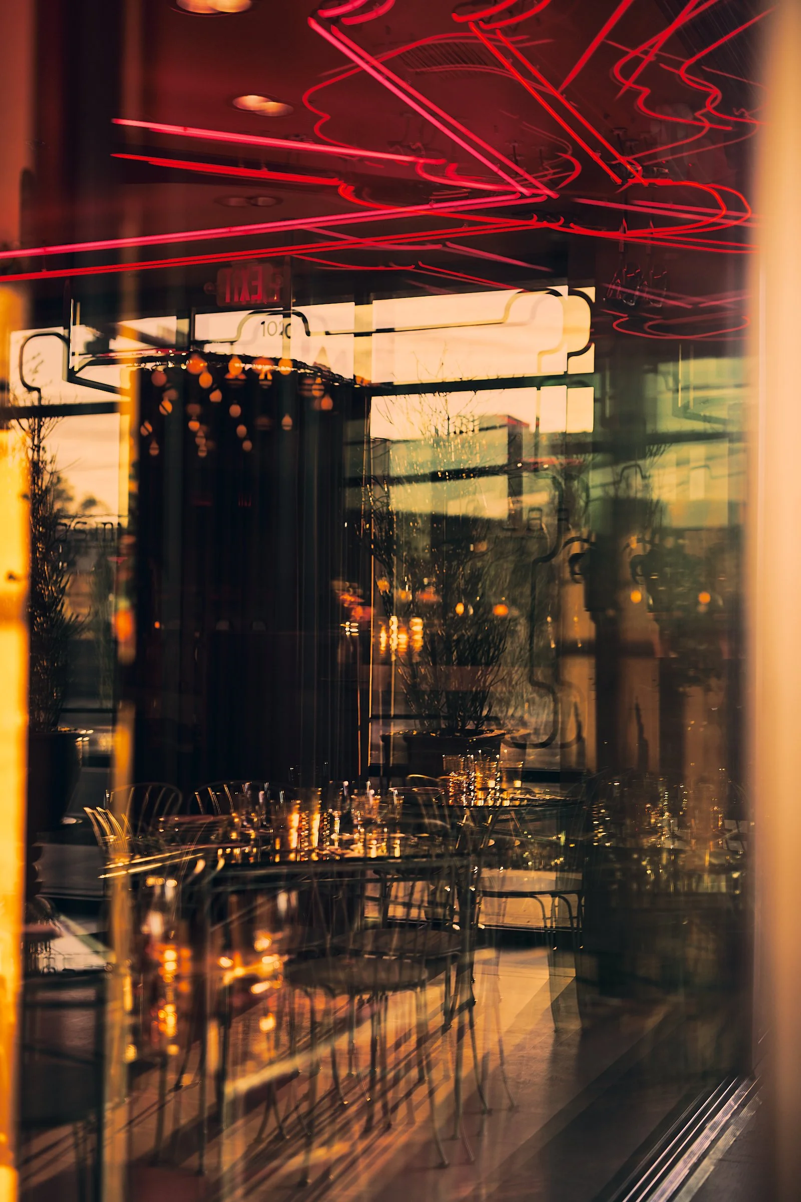 View through a restaurant window showing neon lights inside and reflections of buildings outside at sunset.