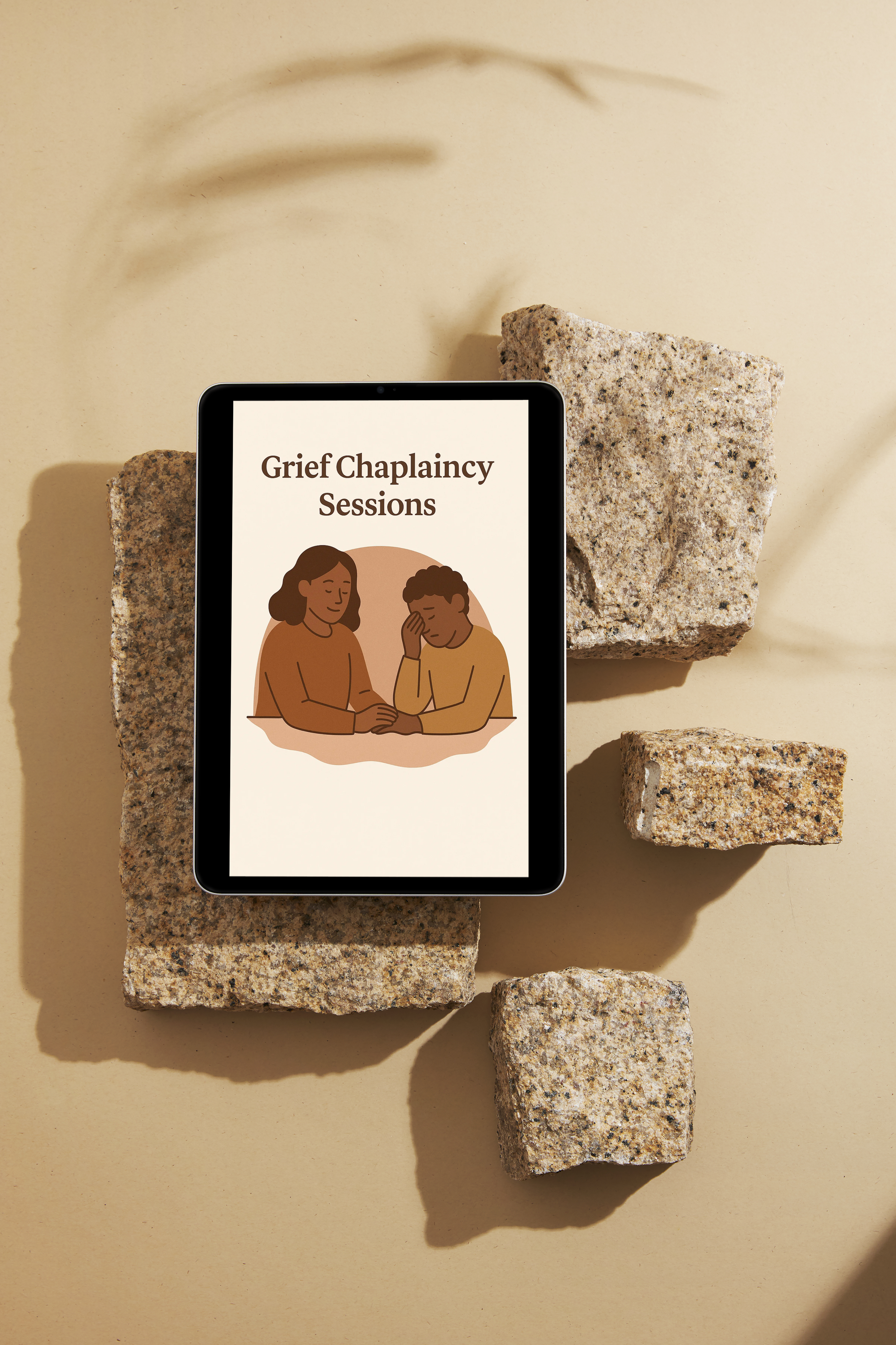 Tablet displaying an illustration with two people in a comforting pose and text that reads "Grief Chaplaincy Sessions," placed on stones with shadowed background.