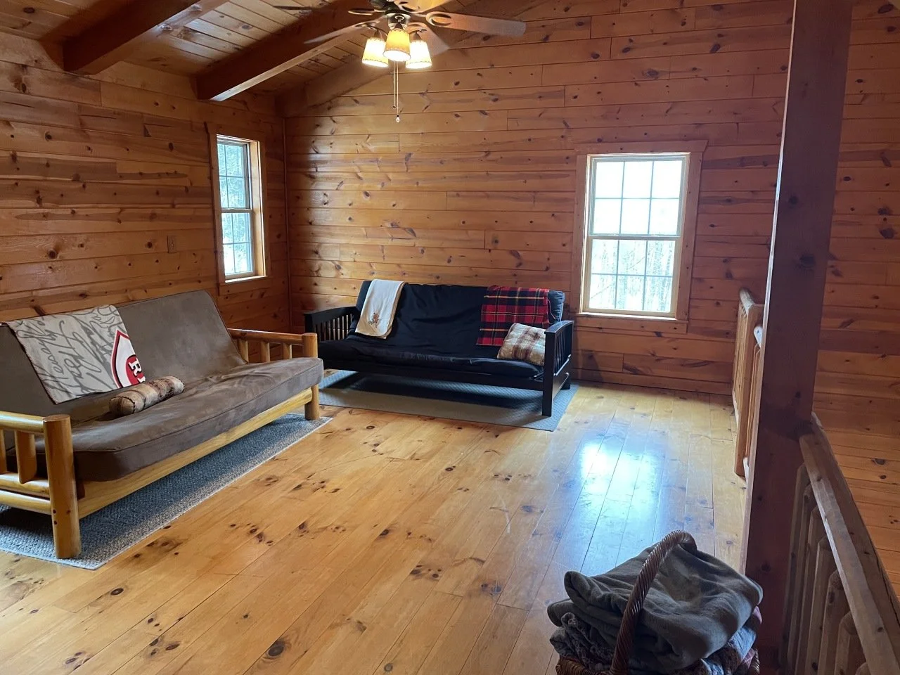 A cozy sitting area with wooden walls, a ceiling fan with lights, and two windows. There are two couches: one with a beige cushion and sports-themed pillows, and another black wooden frame with a blanket and pillows. A small rug partially covers the