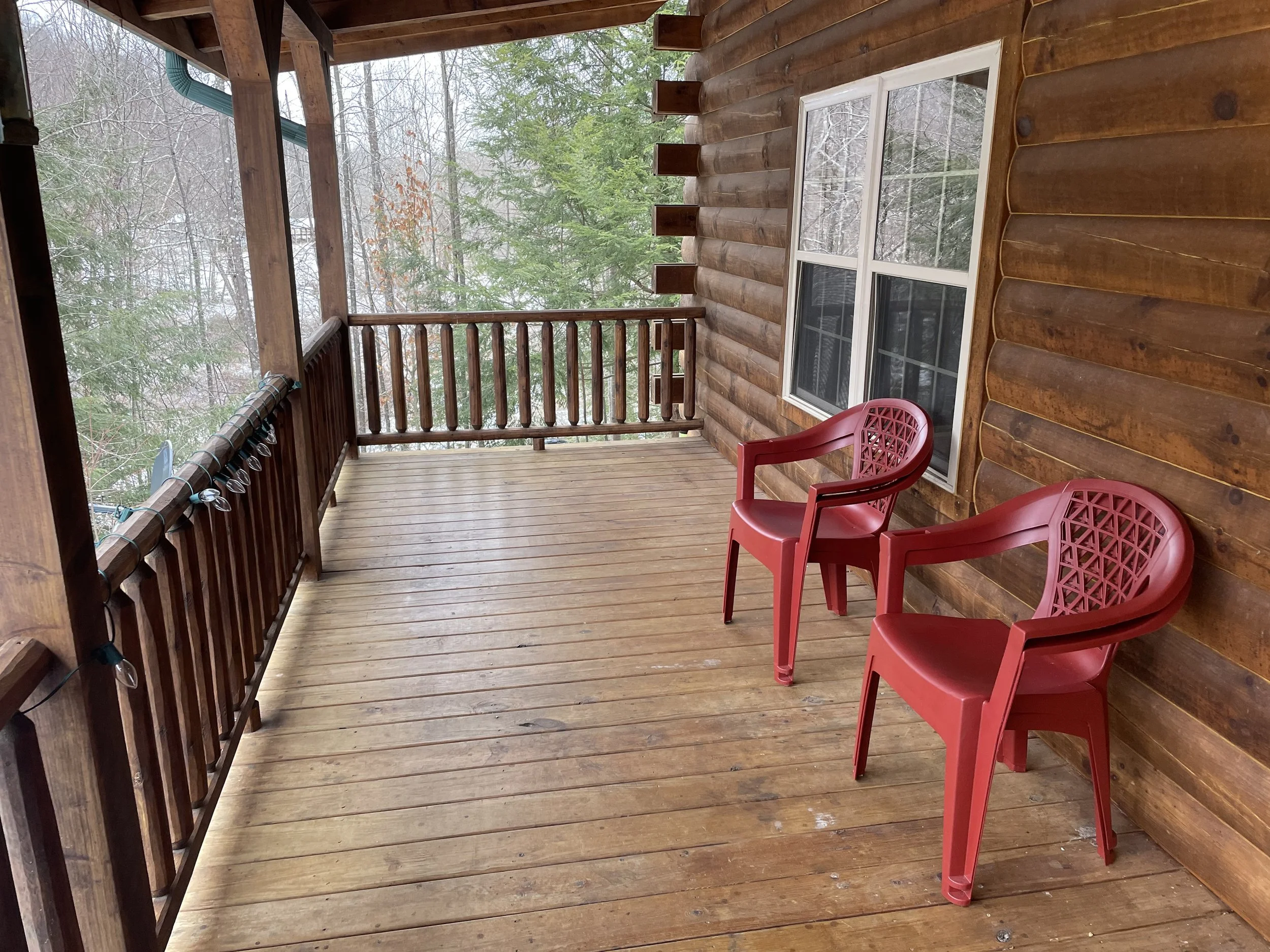 A wooden porch with two red plastic chairs, a log cabin wall, a window, and a wooded background with trees and snow.