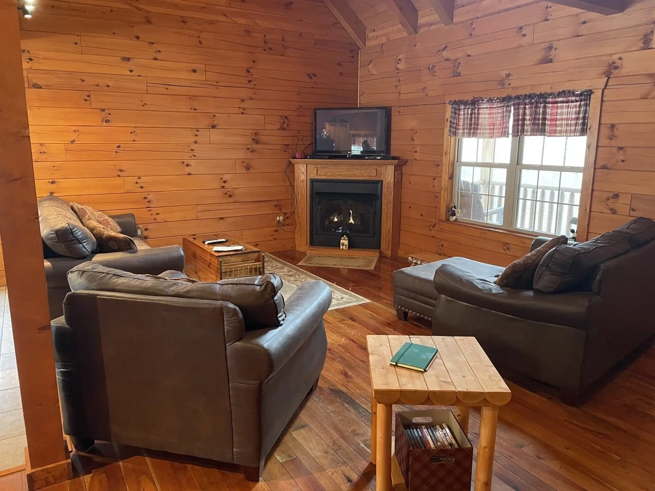 A cozy living room with wooden walls, a fireplace with a flat-screen TV on top, and multiple leather sofas. There is a window with plaid curtains, a small wooden table with a notebook and DVDs, and a rug near the fireplace.