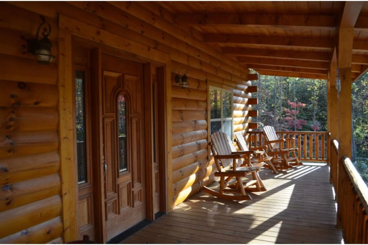 Wooden porch with three Adirondack rocking chairs overlooking a wooded area with trees and colorful fall foliage.