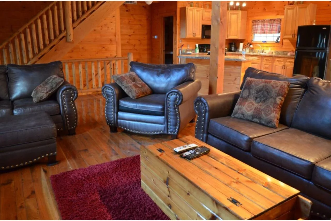 Living room with leather sofas and armchair, wooden coffee table, in an open-plan log cabin with kitchen in background.