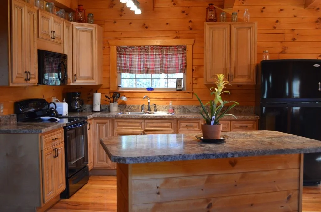 Rustic kitchen with wooden cabinets, granite countertops, a black refrigerator, and a small window with a red plaid curtain
