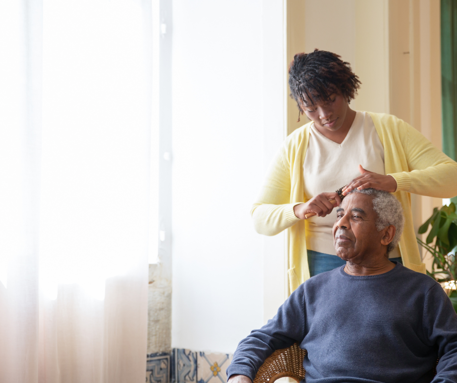 A young home attendants woman styles an elderly man's gray hair with a brush or comb in a bright room with sunlight coming through sheer curtains.