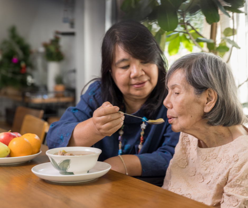 A woman assisting an elderly woman in eating soup at a wooden table with a bowl, a plate of fruit, and a cozy, plant-filled kitchen in the background.