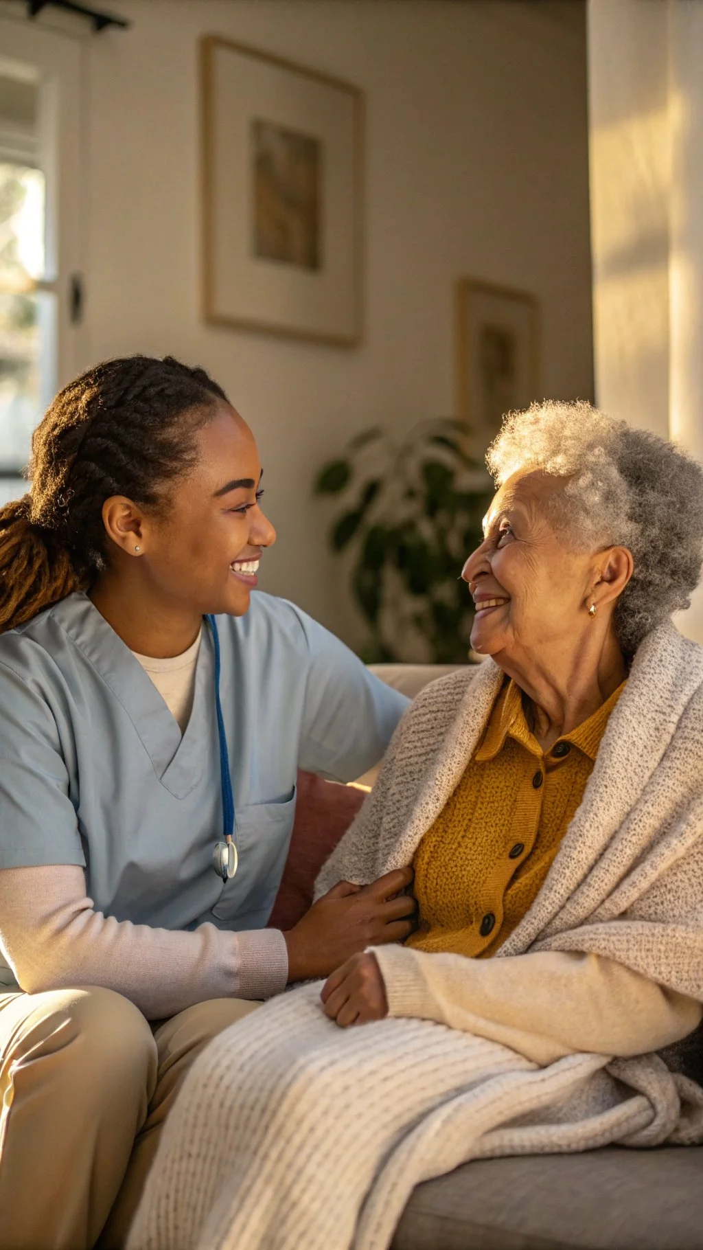 A young female nurse or caregiver smiling and talking with an elderly woman sitting on a couch in a cozy living room, illuminated by warm sunlight.