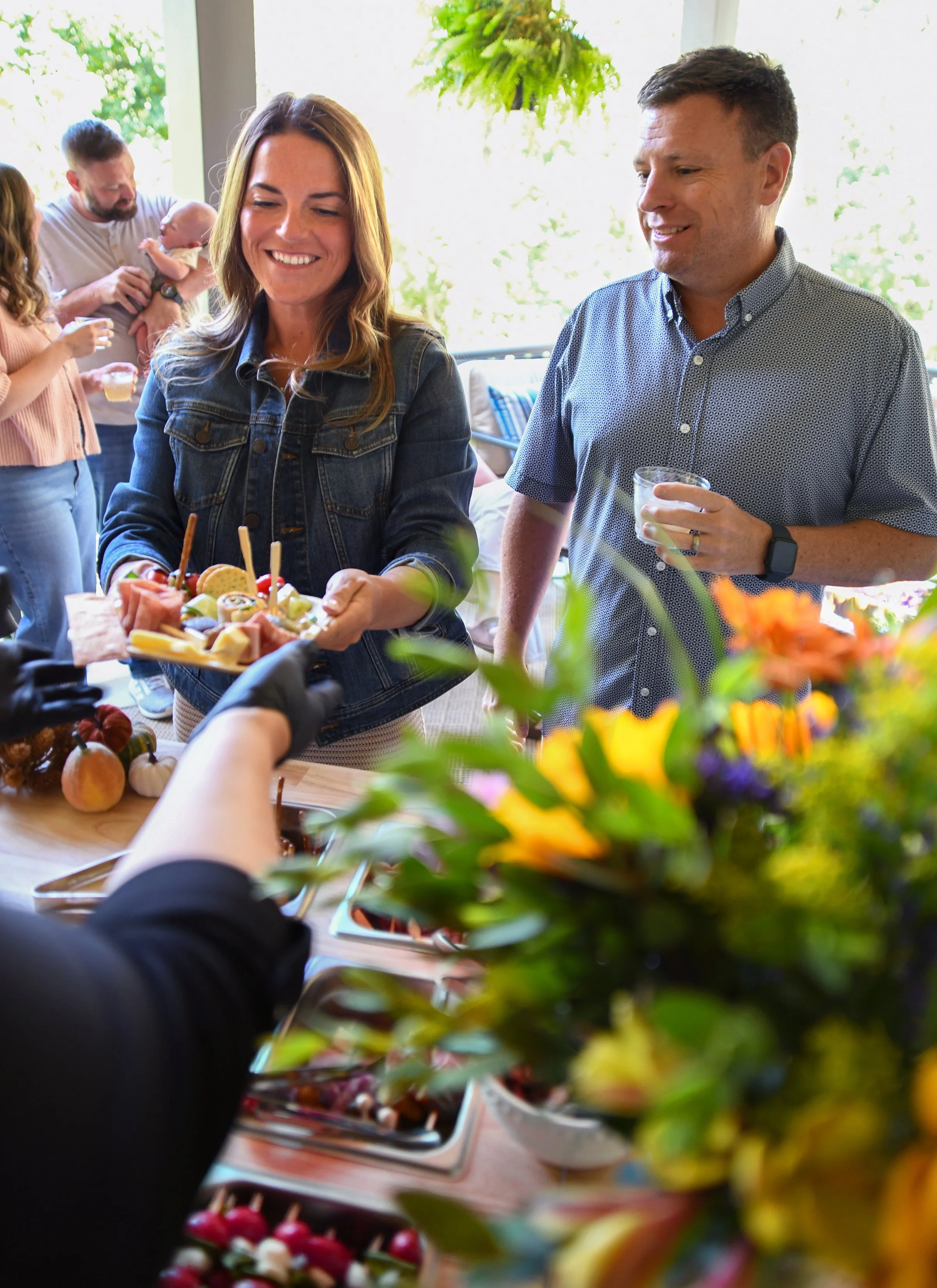 A woman is holding a plate of food at a gathering, smiling. A man next to her is holding a drink and smiling. Other people in the background are socializing, with one holding a baby. The scene is bright.
