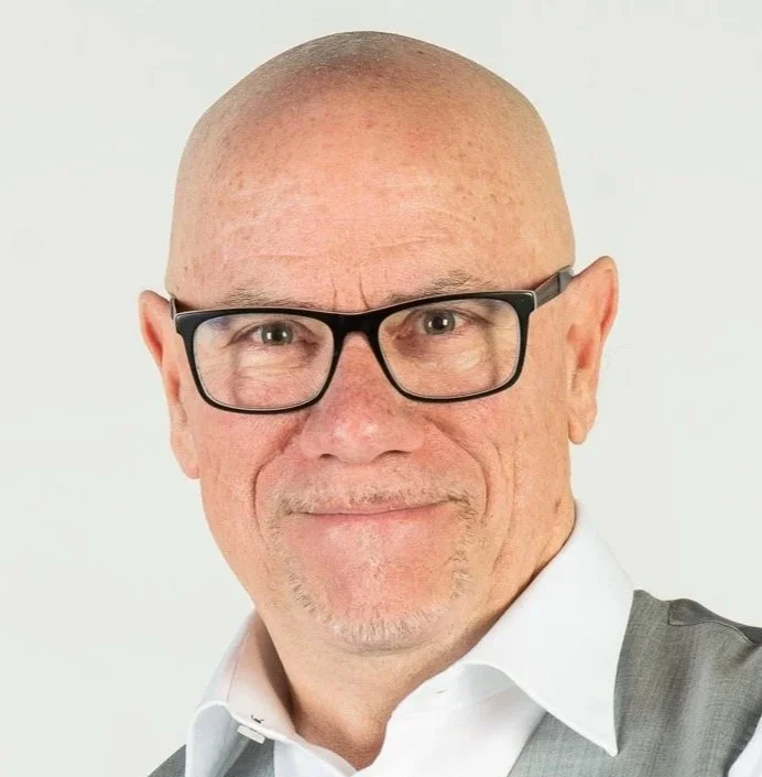 A bald man with glasses smiling, wearing a white shirt and a gray vest, against a plain white background.