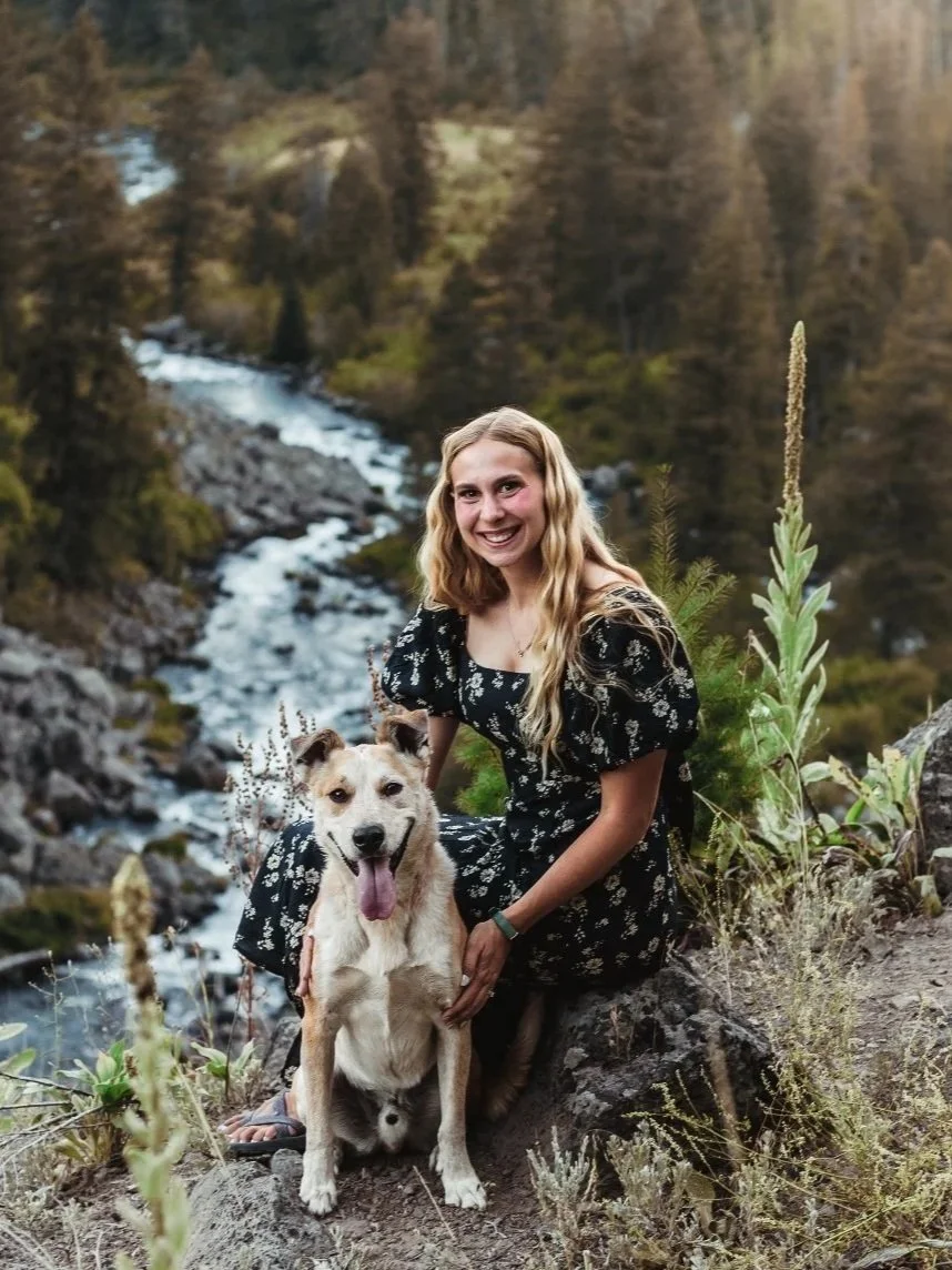 A woman with long blonde hair in a black floral dress sitting next to a tan and white dog with a pink tongue amid scenic nature with a river and forested mountains in the background.