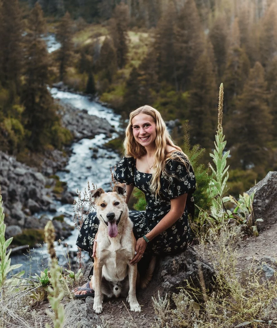 A woman with long blonde hair wearing a black dress with white floral pattern, smiling and sitting on a rock next to a tan and white dog with its tongue out, in a mountainous outdoor setting with trees and a flowing river in the background.