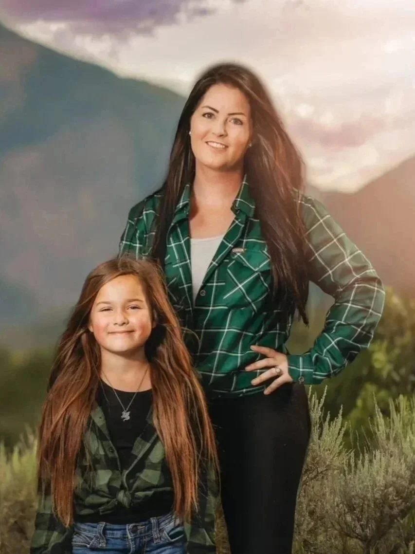 A woman and a girl standing outdoors in a scenic mountainous area, both wearing green plaid shirts, with mountains and a cloudy sky in the background.