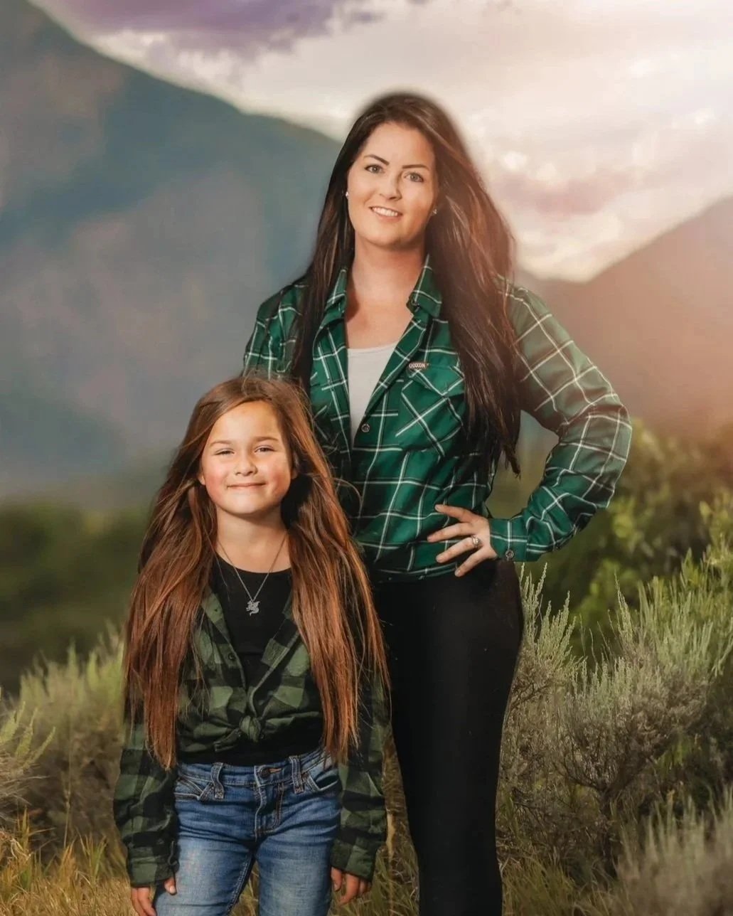 A woman and a young girl standing outdoors in front of a mountain landscape, both wearing green plaid shirts, smiling at the camera.