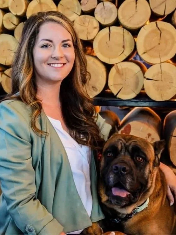 A smiling woman with long wavy brown hair in a green jacket and white shirt, sitting next to a large brown and black dog, Cane Corso, with a tan collar, in front of a background of stacked cut logs.