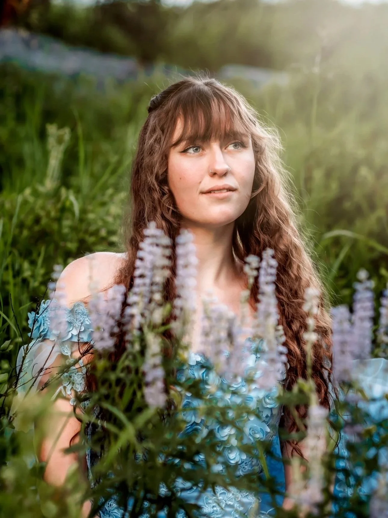 A young woman with long, curly brown hair, wearing a blue dress with floral detail, sitting in a lush green outdoor setting with tall grass and purple flowers, gazing thoughtfully into the distance.